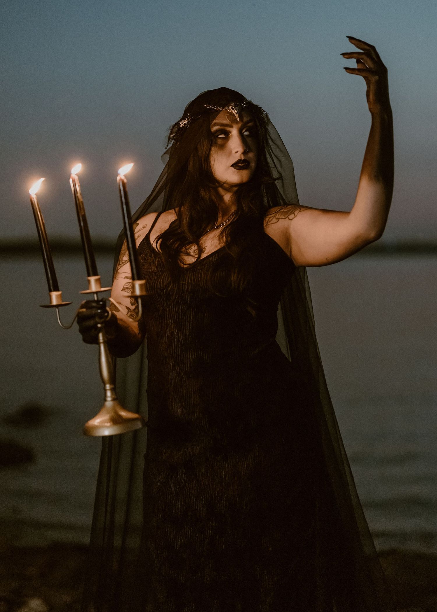 A gothic figure in dark attire poses with a brass candelabra during dusk by a waterfront.