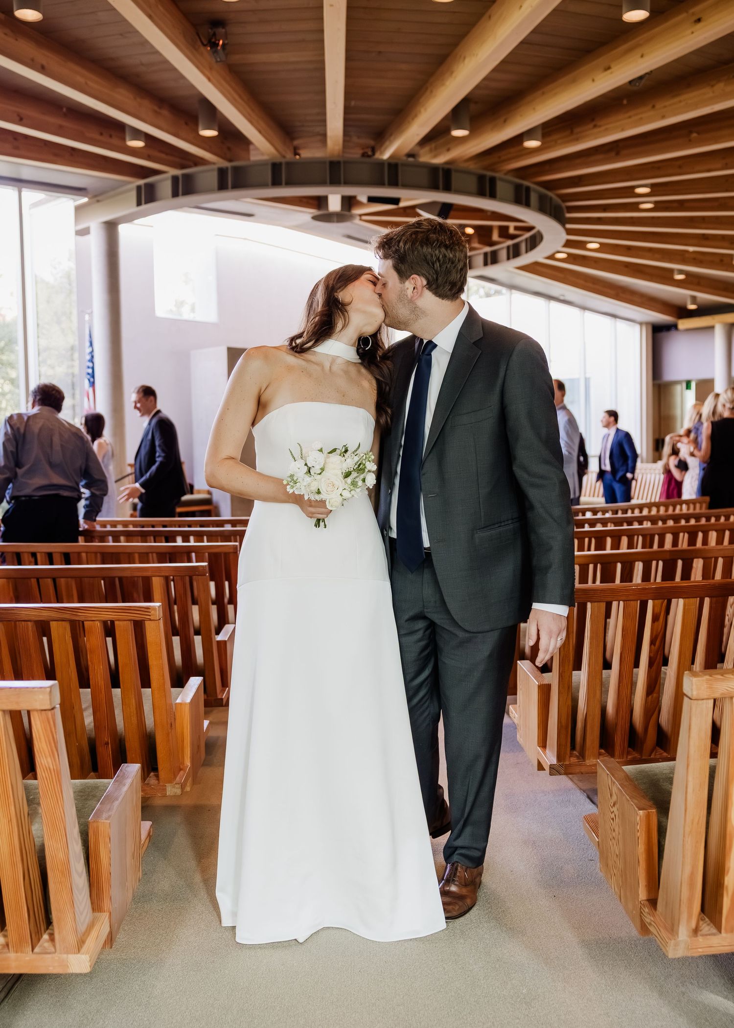 Bride and groom share a first kiss as newlyweds in a modern wooden chapel aisle.