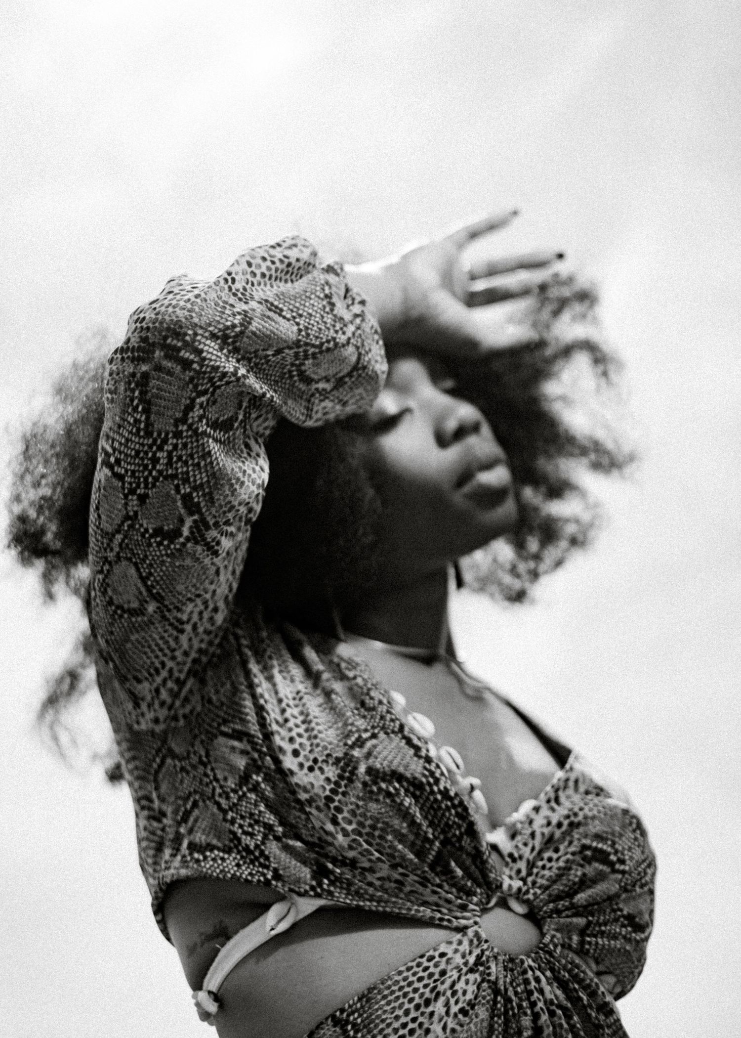 Black and white photo of a woman in snake print top, hair blowing, hand raised to forehead against cloudy sky.