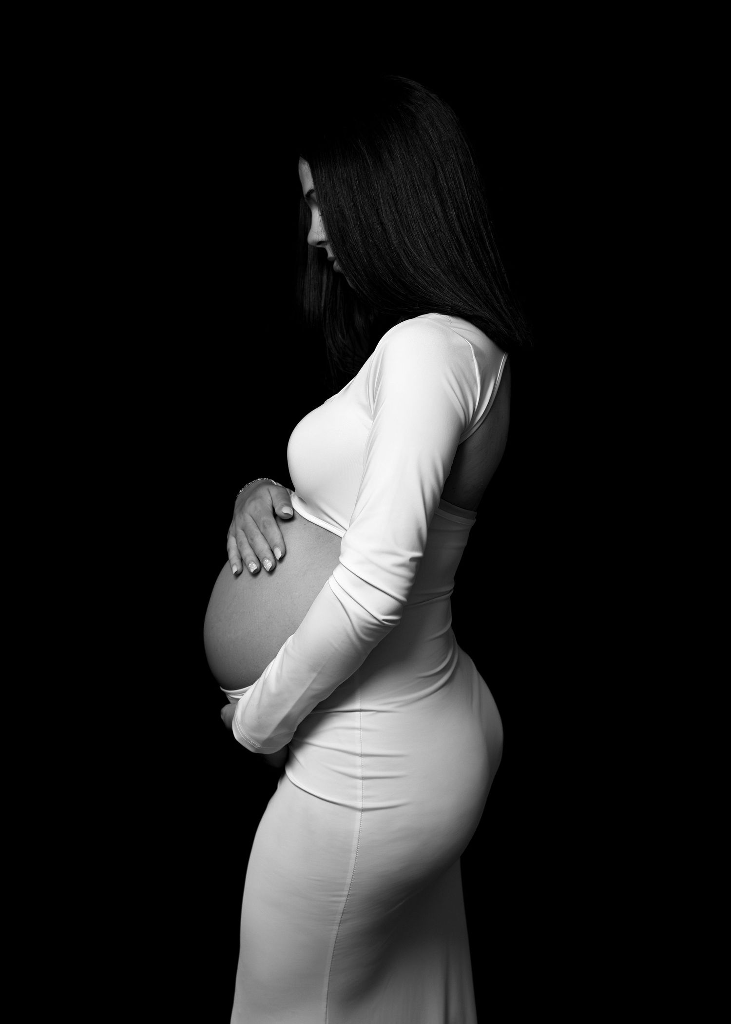 Black and white maternity photo of pregnant woman in white dress cradling baby bump against dark background.