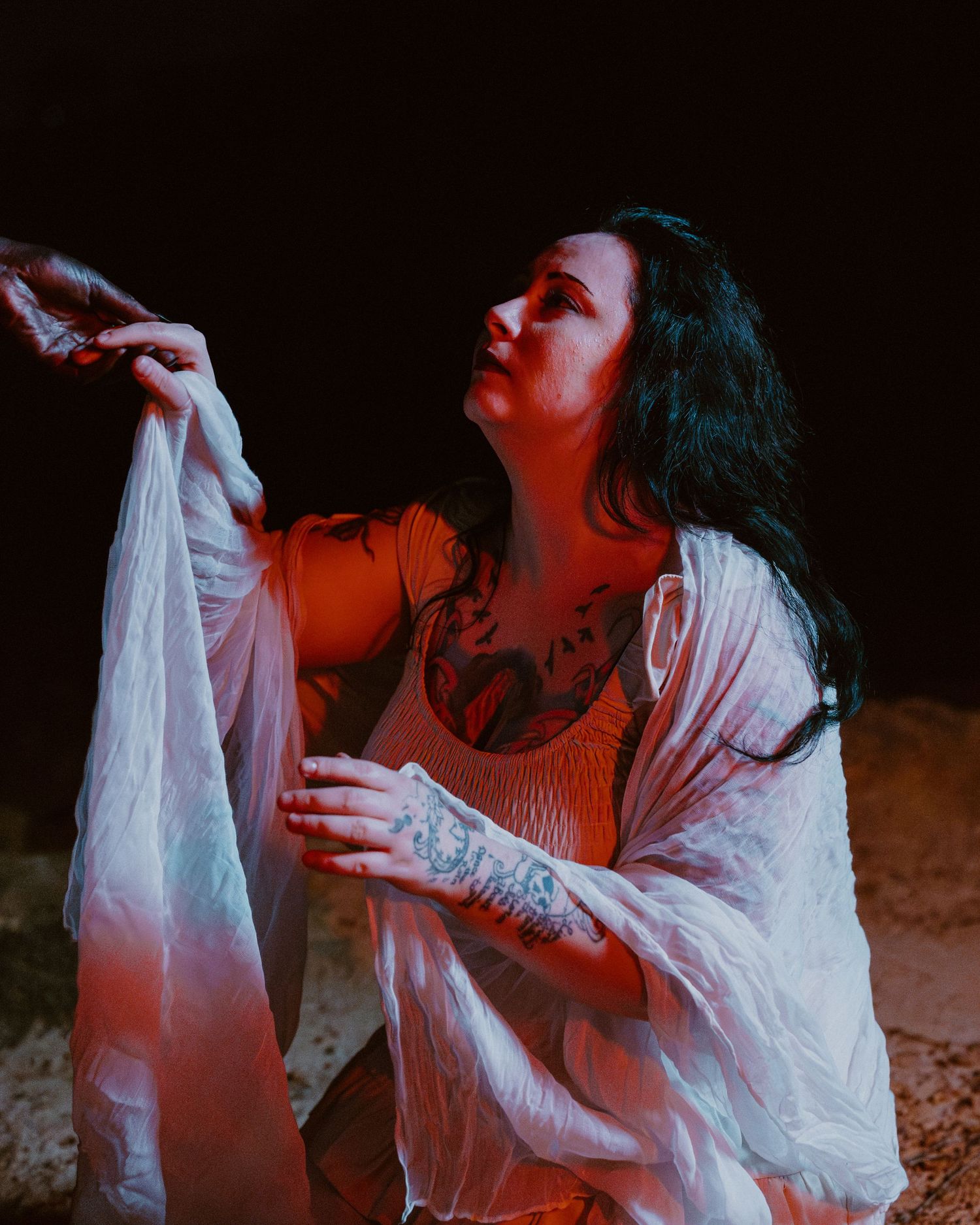 Dramatic portrait with red lighting shows figure in flowing white fabric against dark background.
