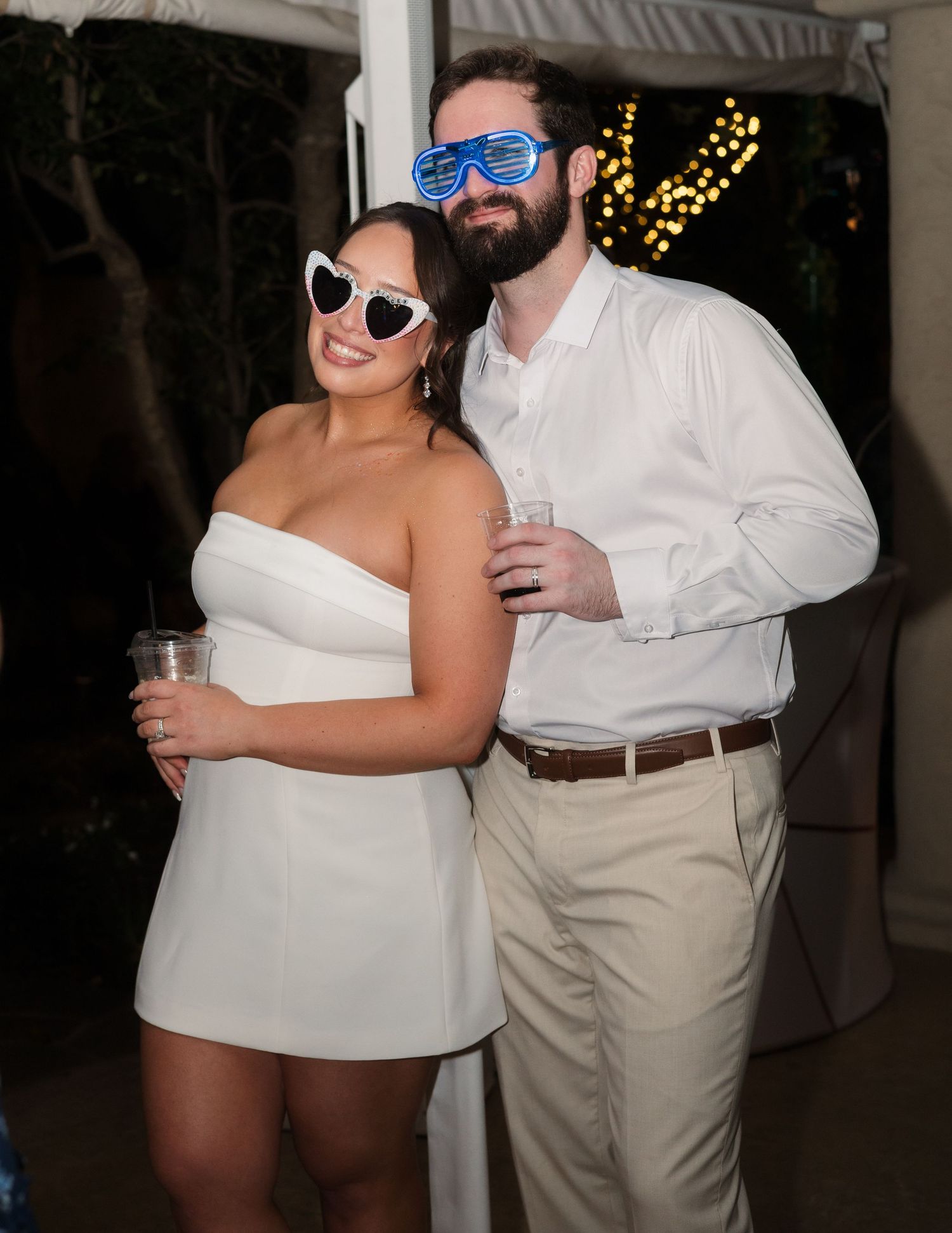 Couple in white outfits wearing novelty sunglasses at an outdoor party, holding drinks and smiling.
