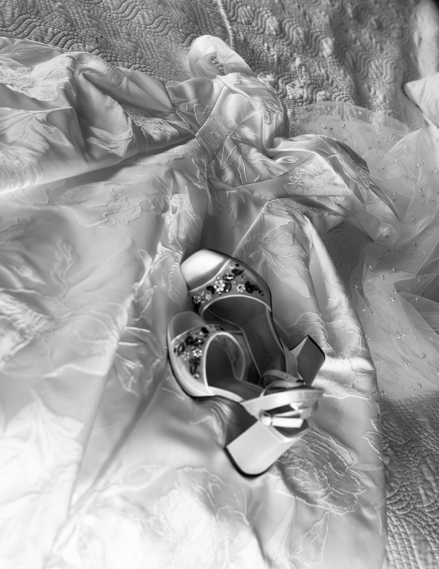 Elegant white bridal heels with crystal embellishments resting on a flowing wedding dress, black and white photo.