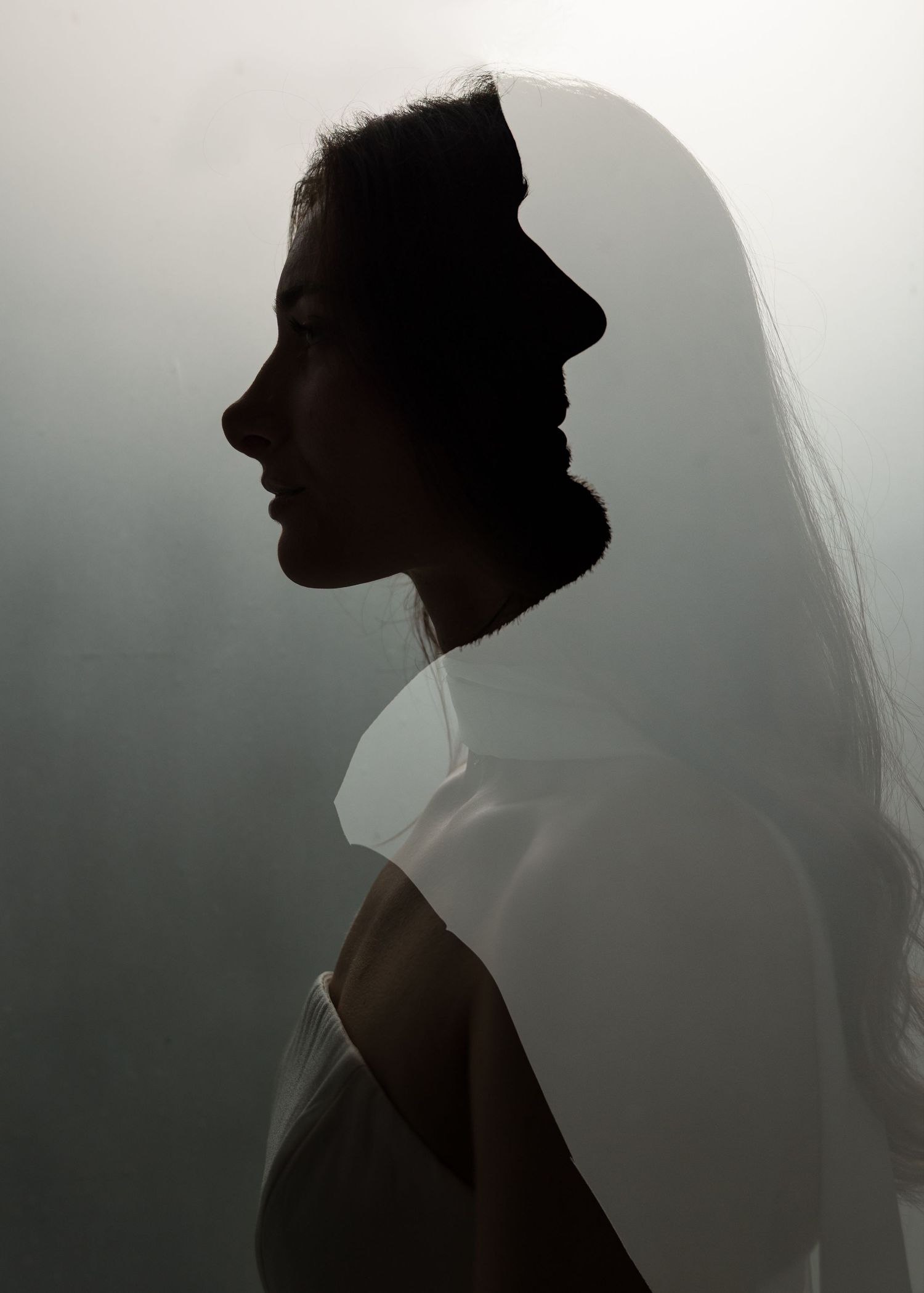 Silhouette of a bride in profile wearing a flowing veil against a soft gray background.