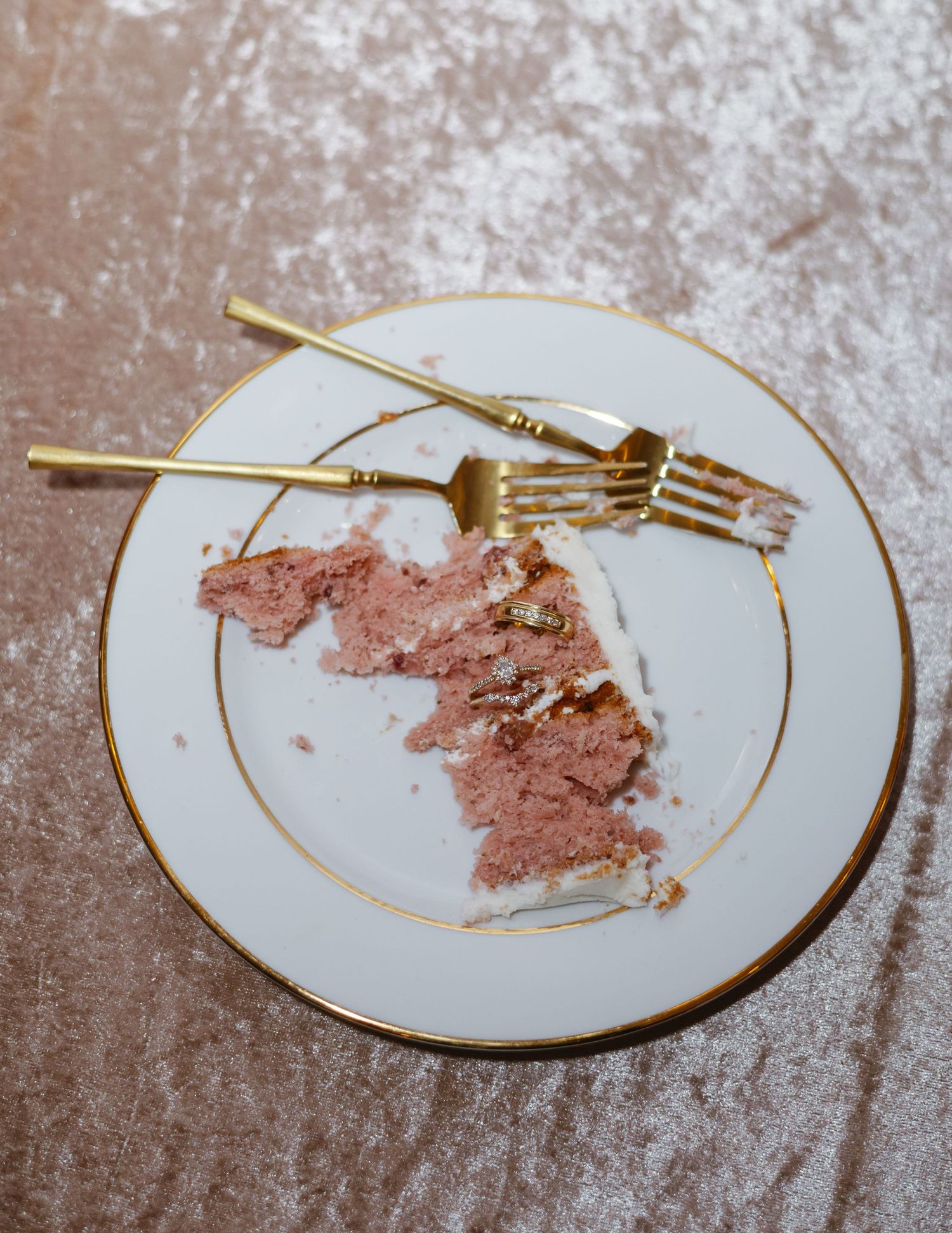 Partially eaten pink cake slice on white gold-rimmed plate with two gold forks on pink velvet surface.