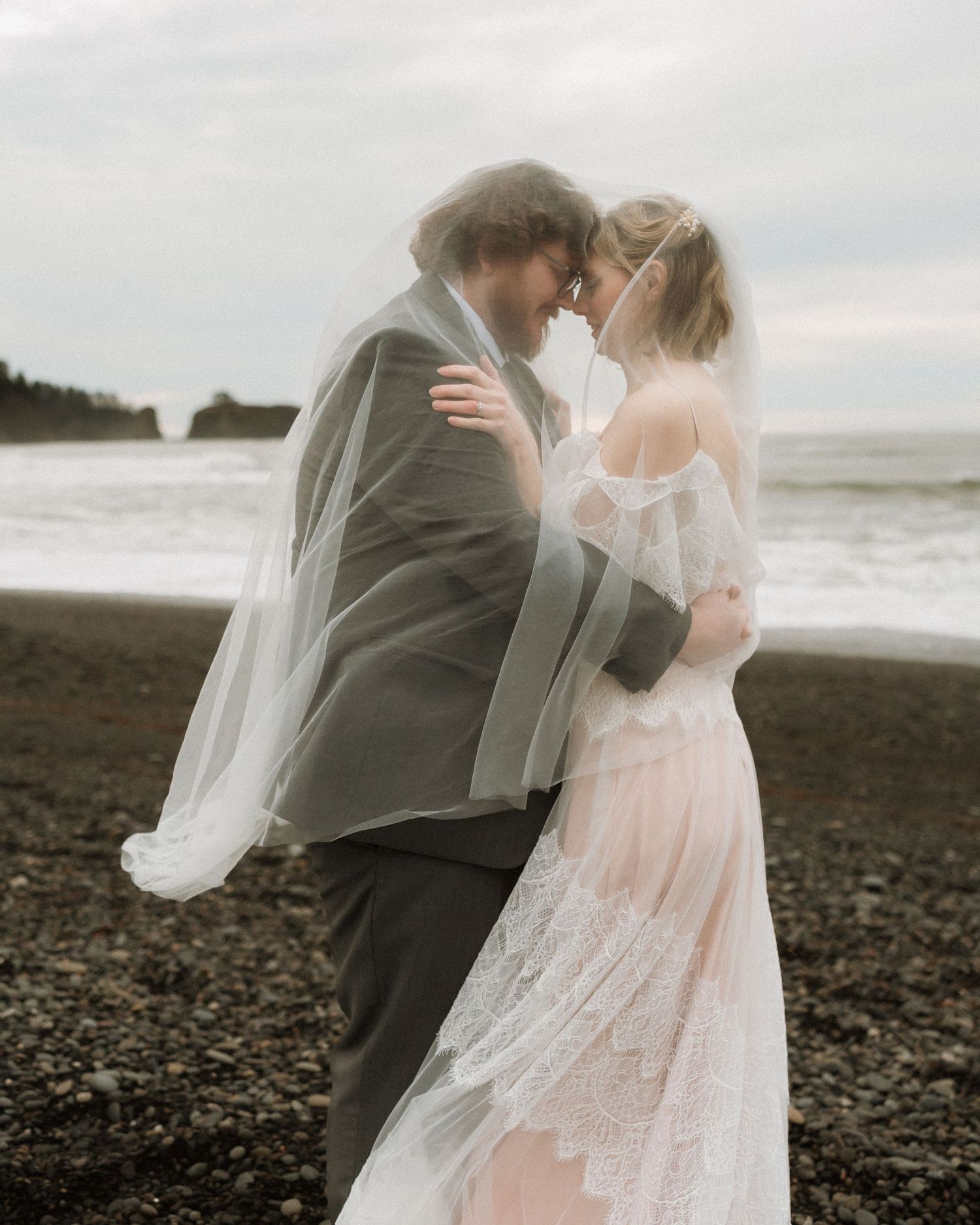 Couple shares romantic moment under flowing veil on black sand beach with ocean waves behind them.