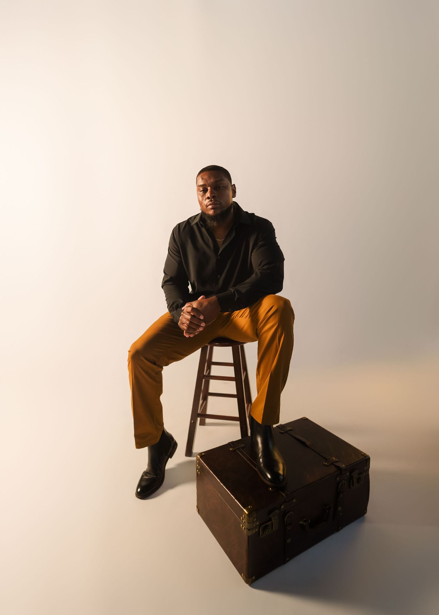 A confident man in a black shirt and mustard trousers sits on a stool over a vintage trunk in a studio.