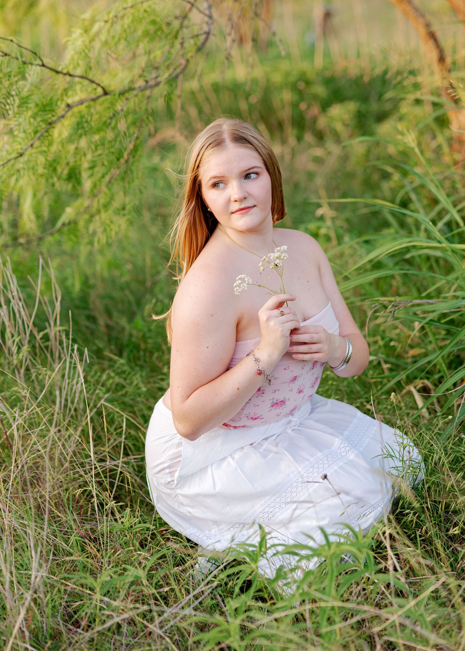 Young girl in white floral dress sitting in tall green grass holding wildflowers during golden hour outdoor photo session.