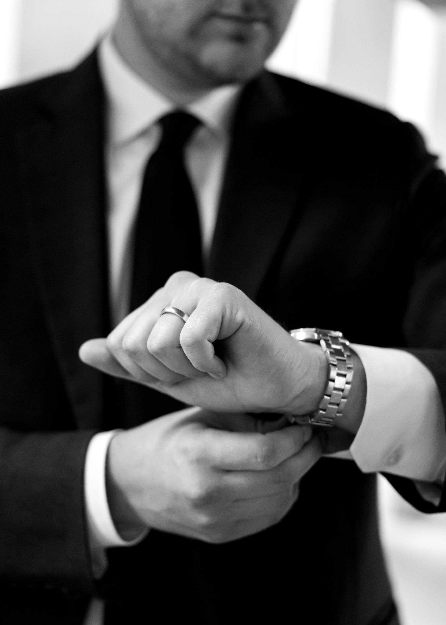 Groom adjusting cufflinks in black suit with watch and wedding ring in black and white photo.