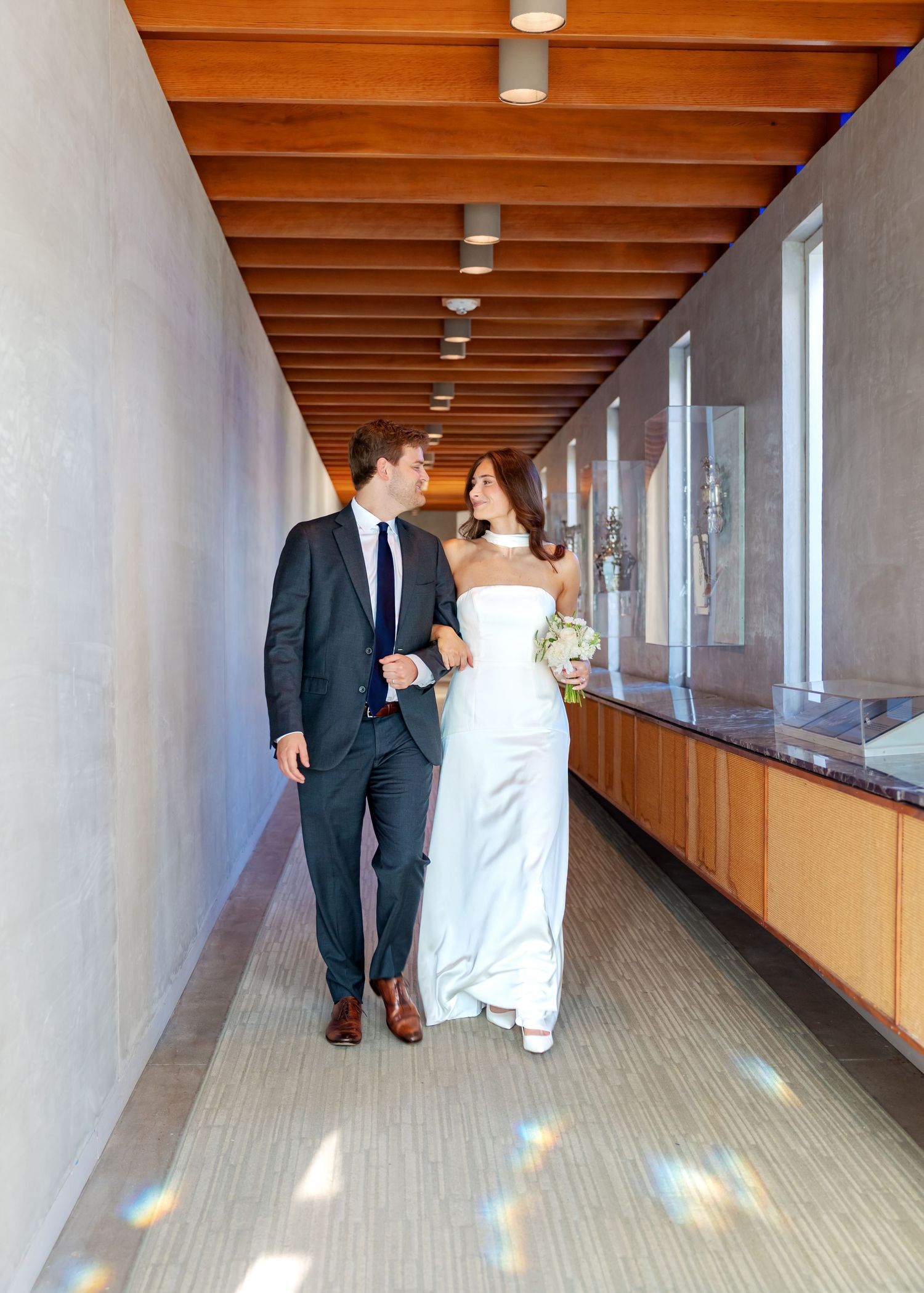 Bride and groom walking together in sunlit wooden hallway, sharing a loving glance.