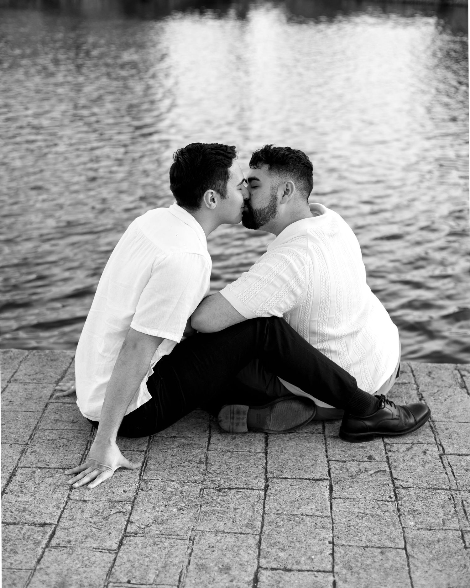 Two men sharing a romantic kiss while sitting on cobblestone pavement by a reflective lake in black and white.