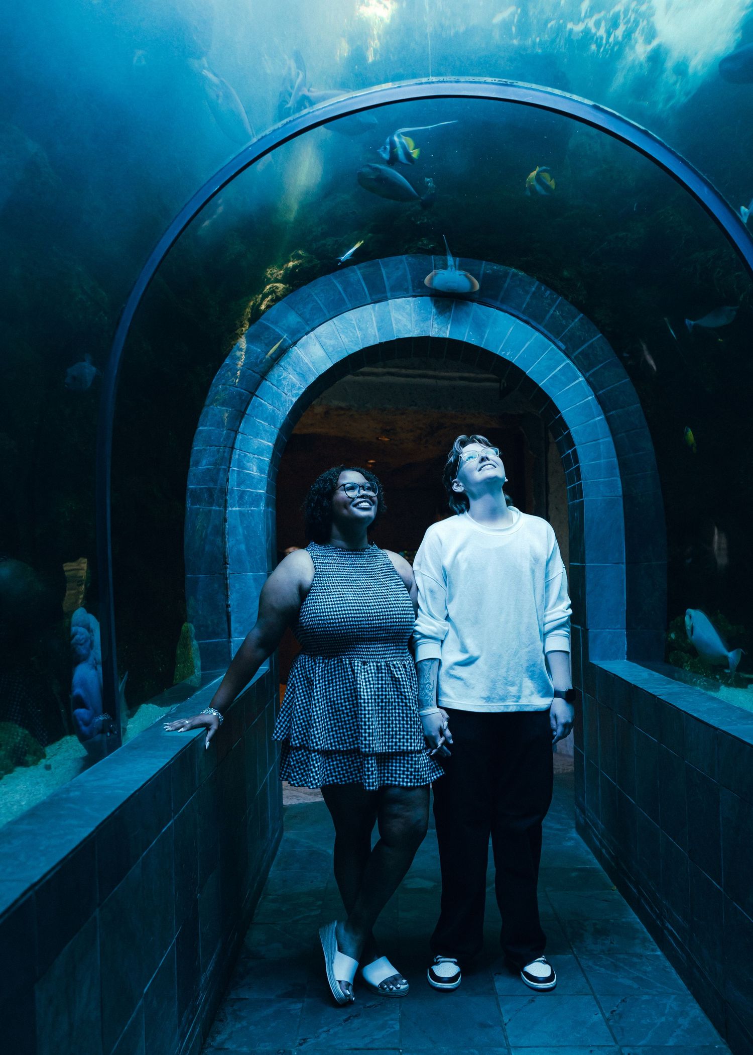 Couple smiling and looking up in an underwater aquarium tunnel surrounded by fish and marine life.