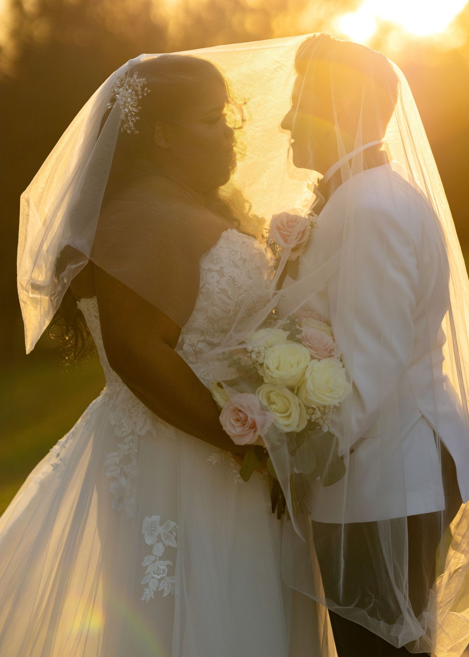 Bride and groom share romantic sunset moment under wedding veil, bride holding pink and white rose bouquet.