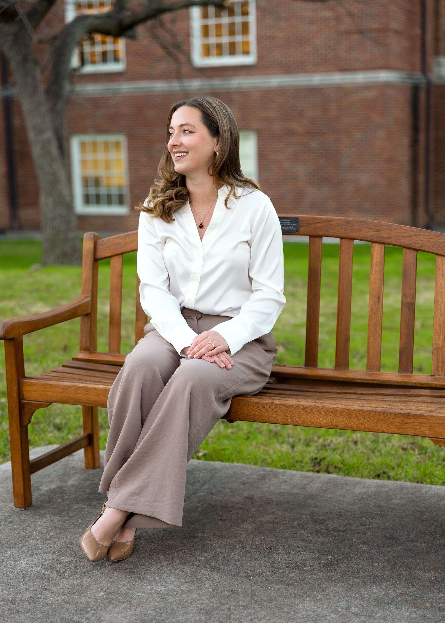 Professional portrait of someone seated on a wooden bench wearing a white blouse and tan pants in front of a brick building.