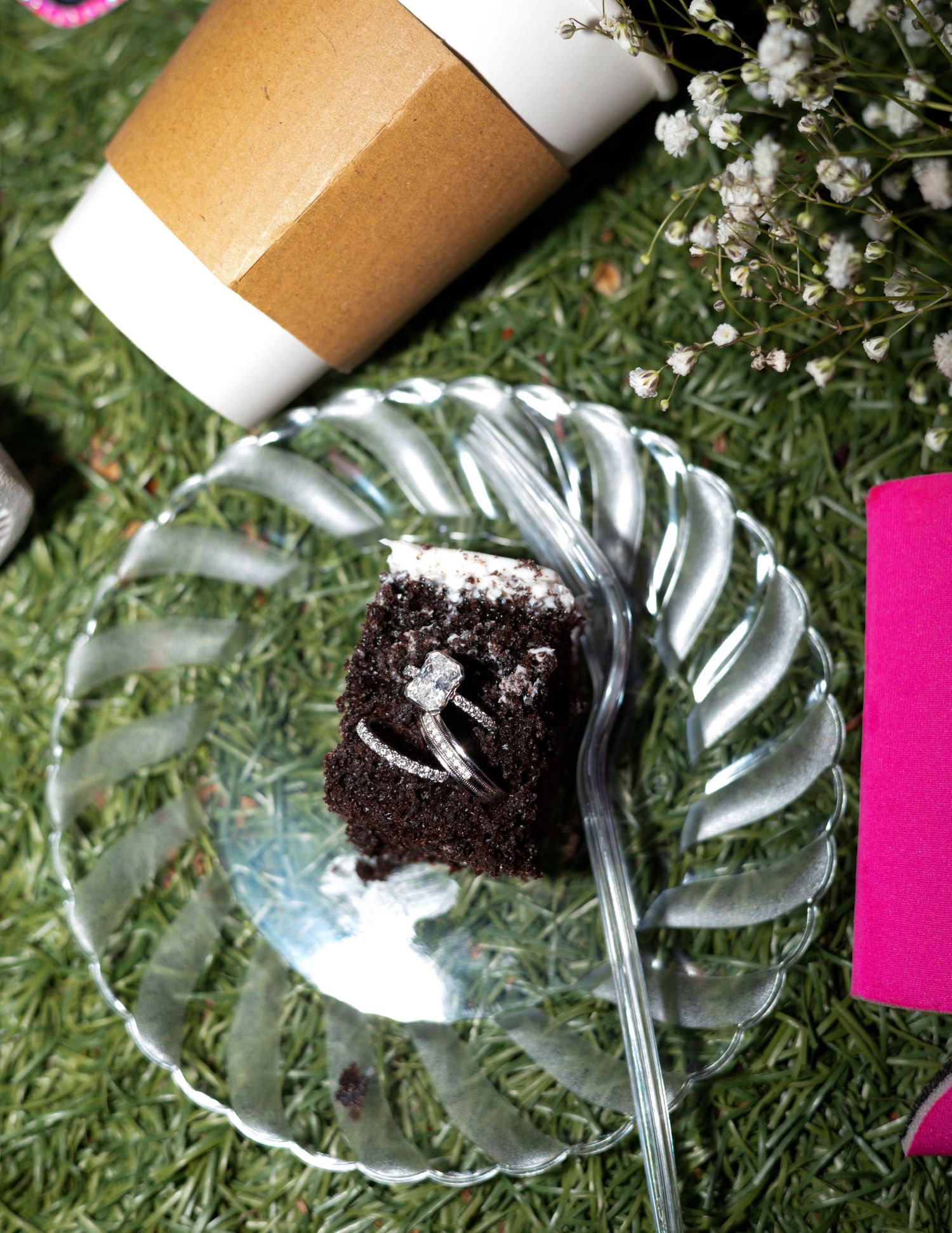 Diamond engagement ring displayed on a chocolate cake slice on a clear plate beside a coffee cup outdoors.