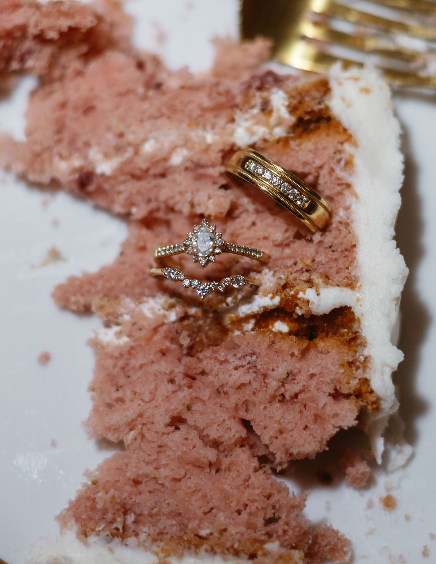 Gold wedding and engagement rings with diamonds displayed on a slice of pink wedding cake with white frosting.