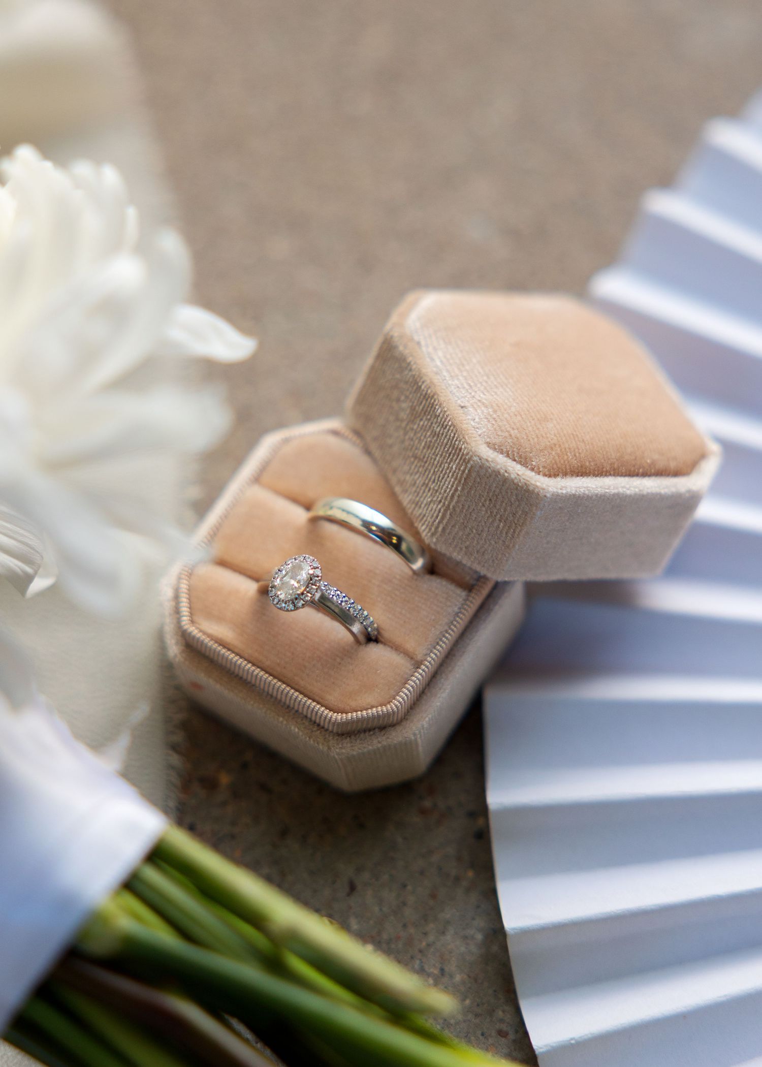 Diamond engagement ring and wedding band displayed in a velvet hexagon ring box beside white flowers and a bouquet.