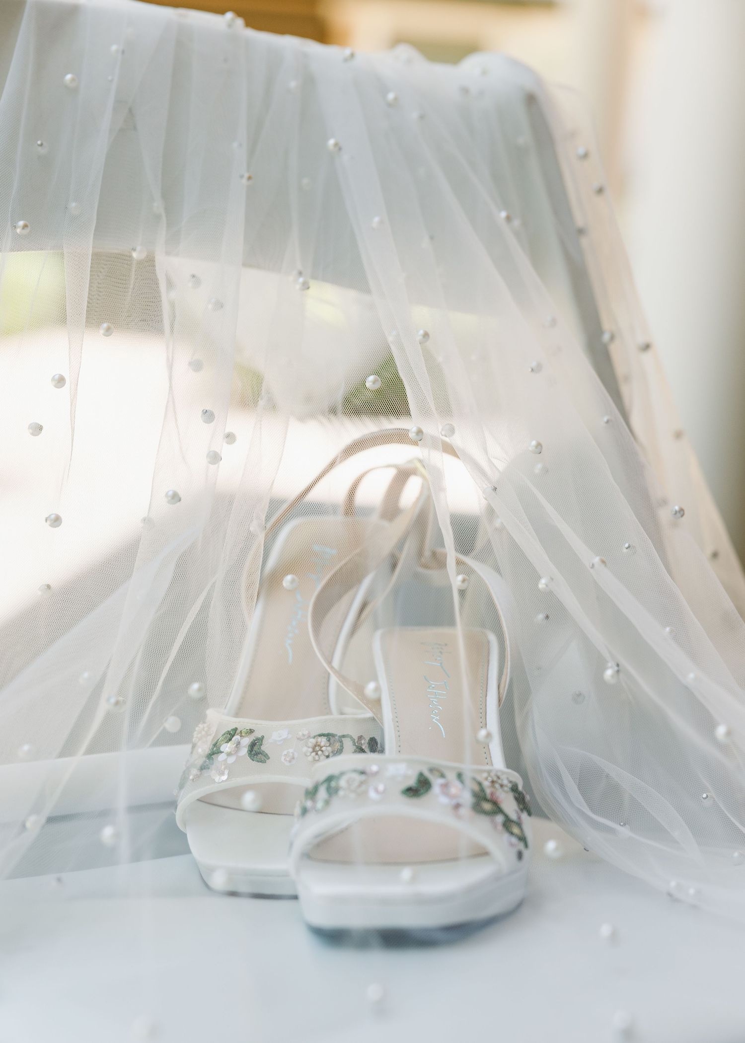 Delicate floral wedding heels displayed against a pearl-embellished bridal veil in soft white tones.