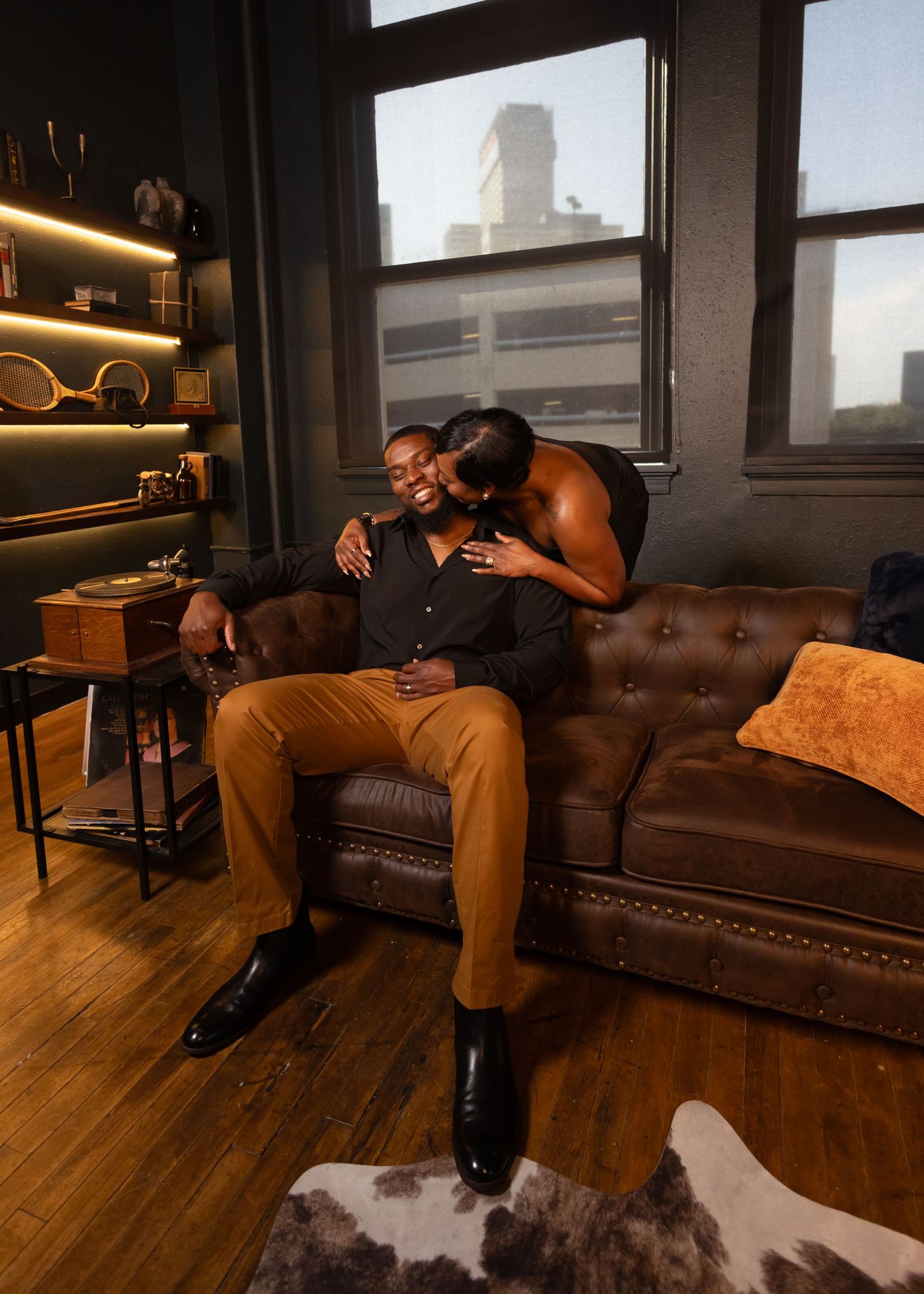 Couple sharing an intimate moment on a leather tufted sofa in a stylish dark loft apartment.
