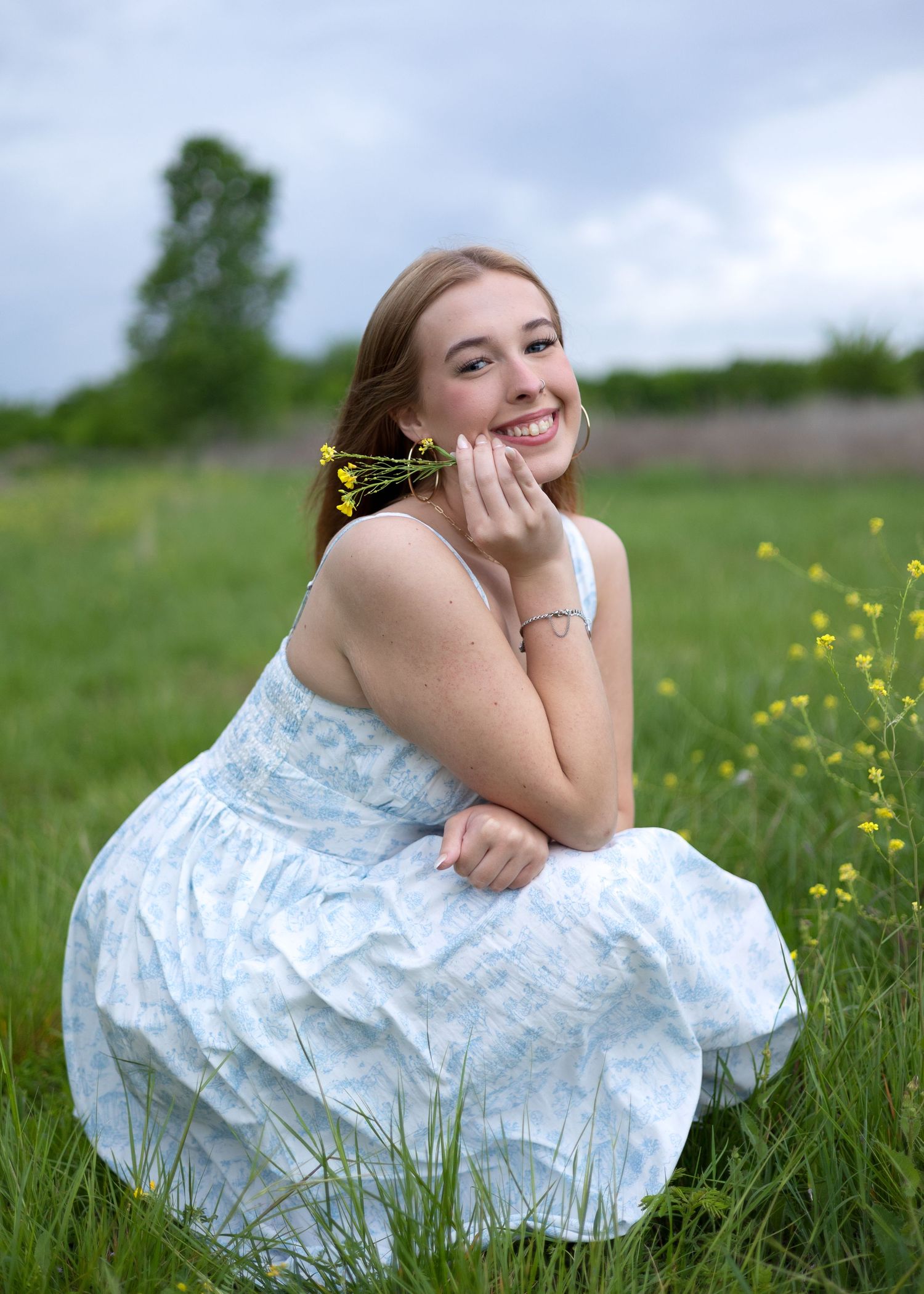 Young woman in a light blue dress sitting in a green field, smiling and holding yellow wildflowers on a cloudy day.