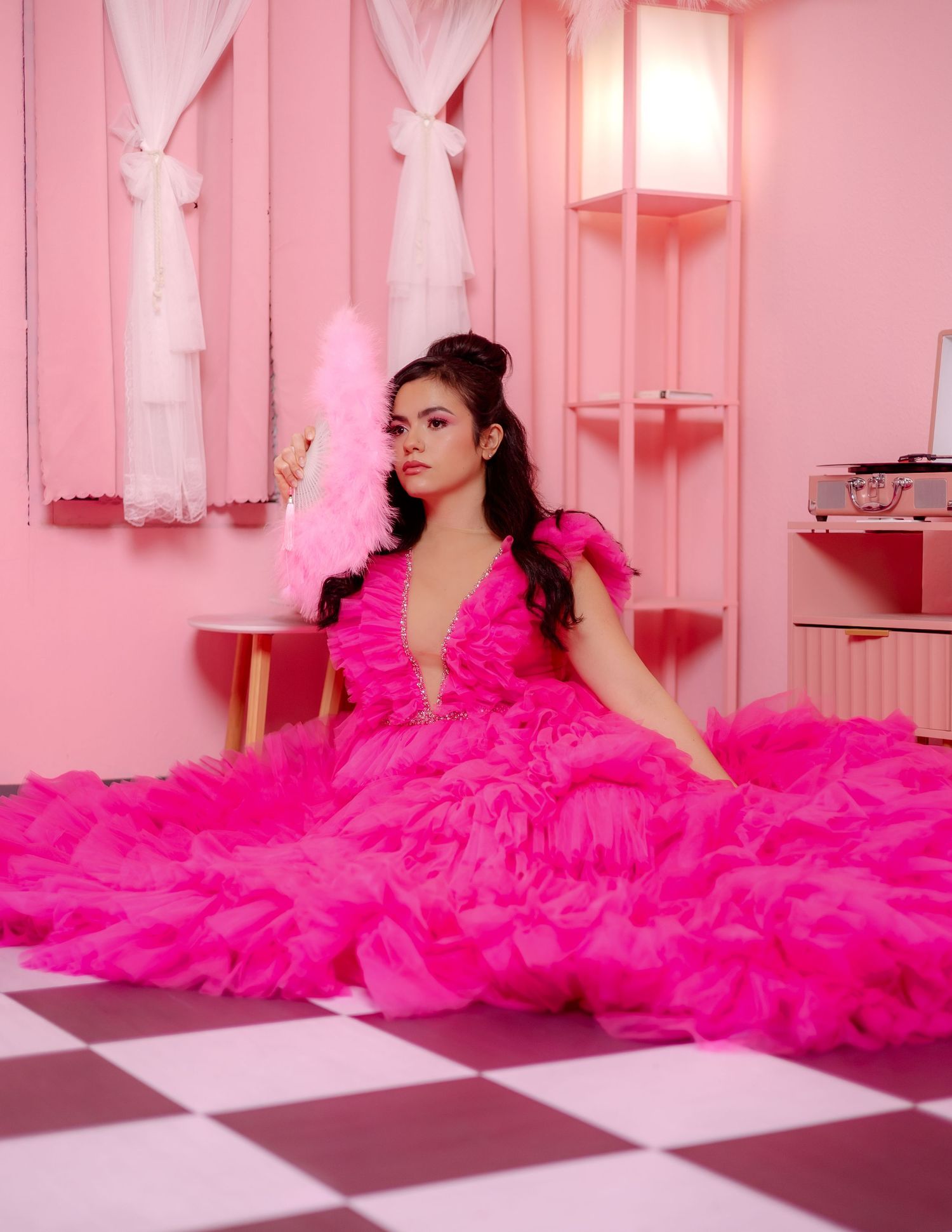 Person lying on checkered floor wearing dramatic flowing hot pink tulle gown in pink-toned studio setting.