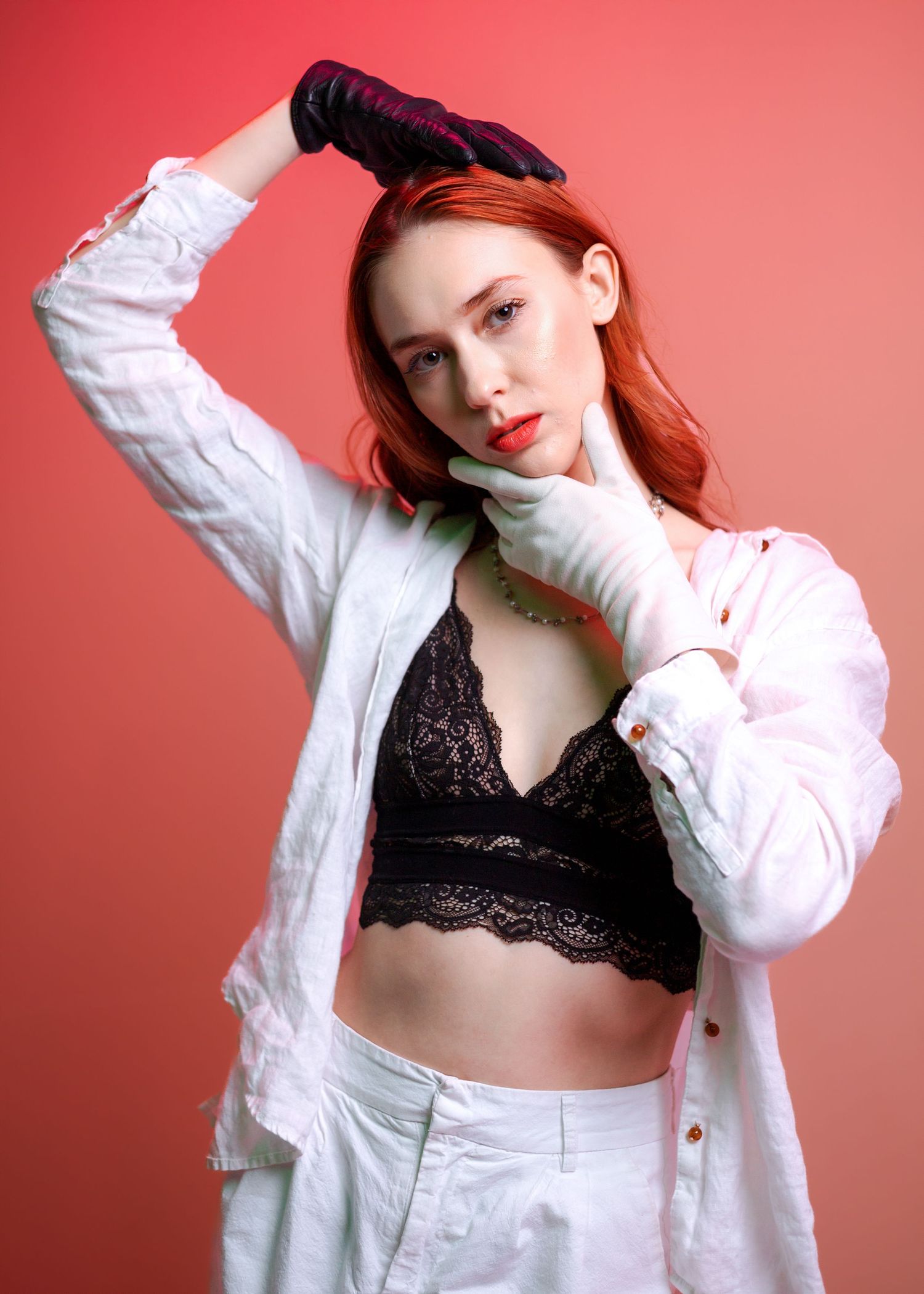 Model in white jacket and black lace bralette poses against coral background with gloved hands.