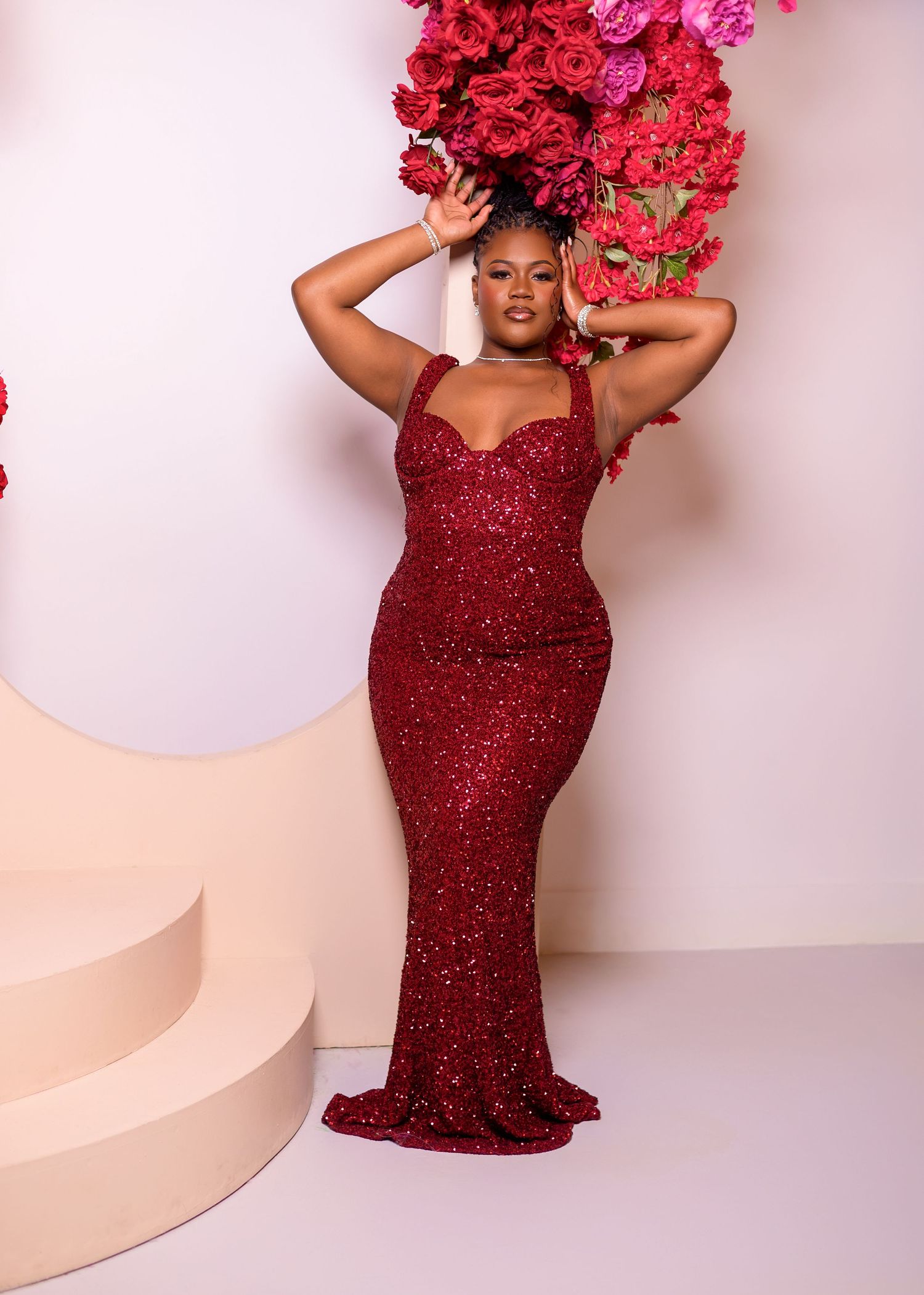A glamorous figure poses in a sparkly burgundy evening gown with bright pink floral decorations against a light backdrop.