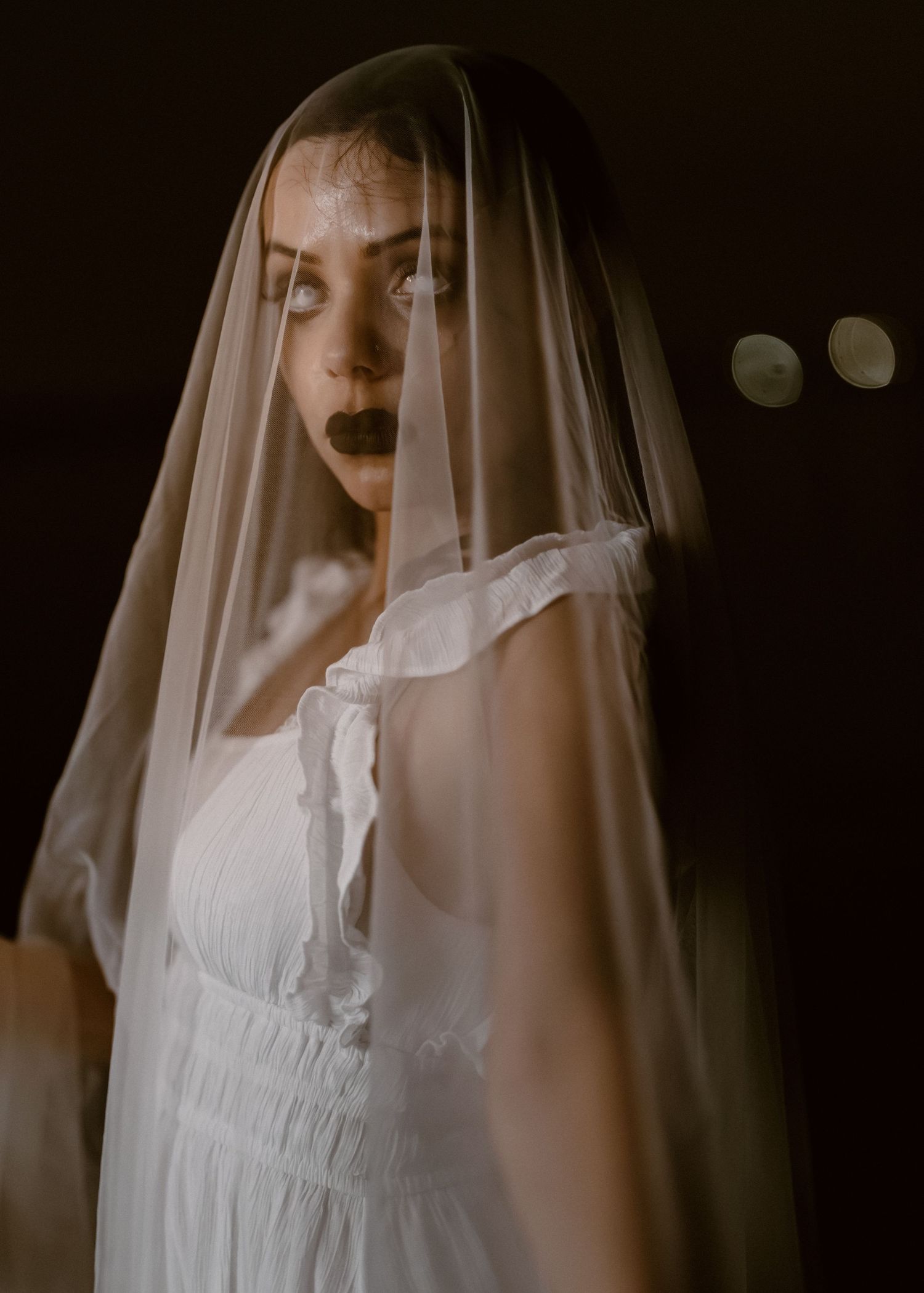 Moody portrait of figure in white dress and veil against dark background with candlelight.