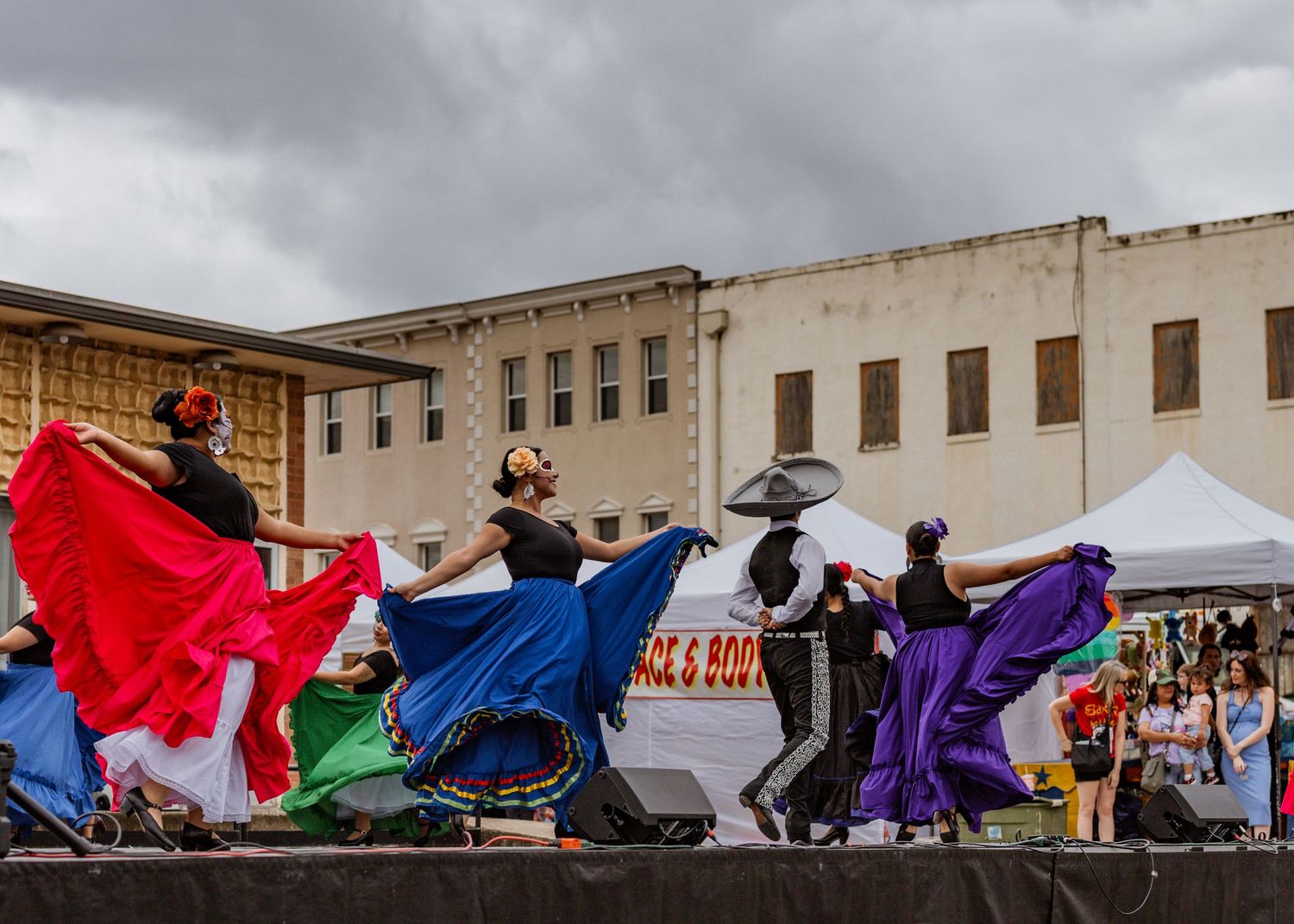 Dancers in flowing colorful costumes perform a choreographed routine against a cloudy sky backdrop.