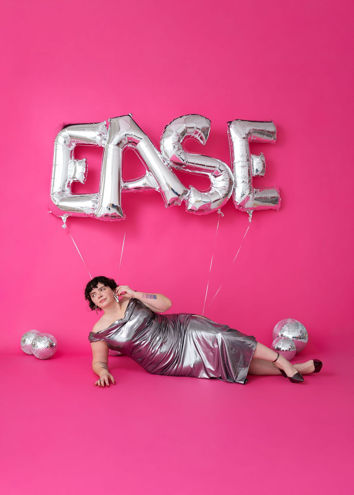 A person in a metallic silver dress poses on a pink background with silver balloon letters spelling EASE.