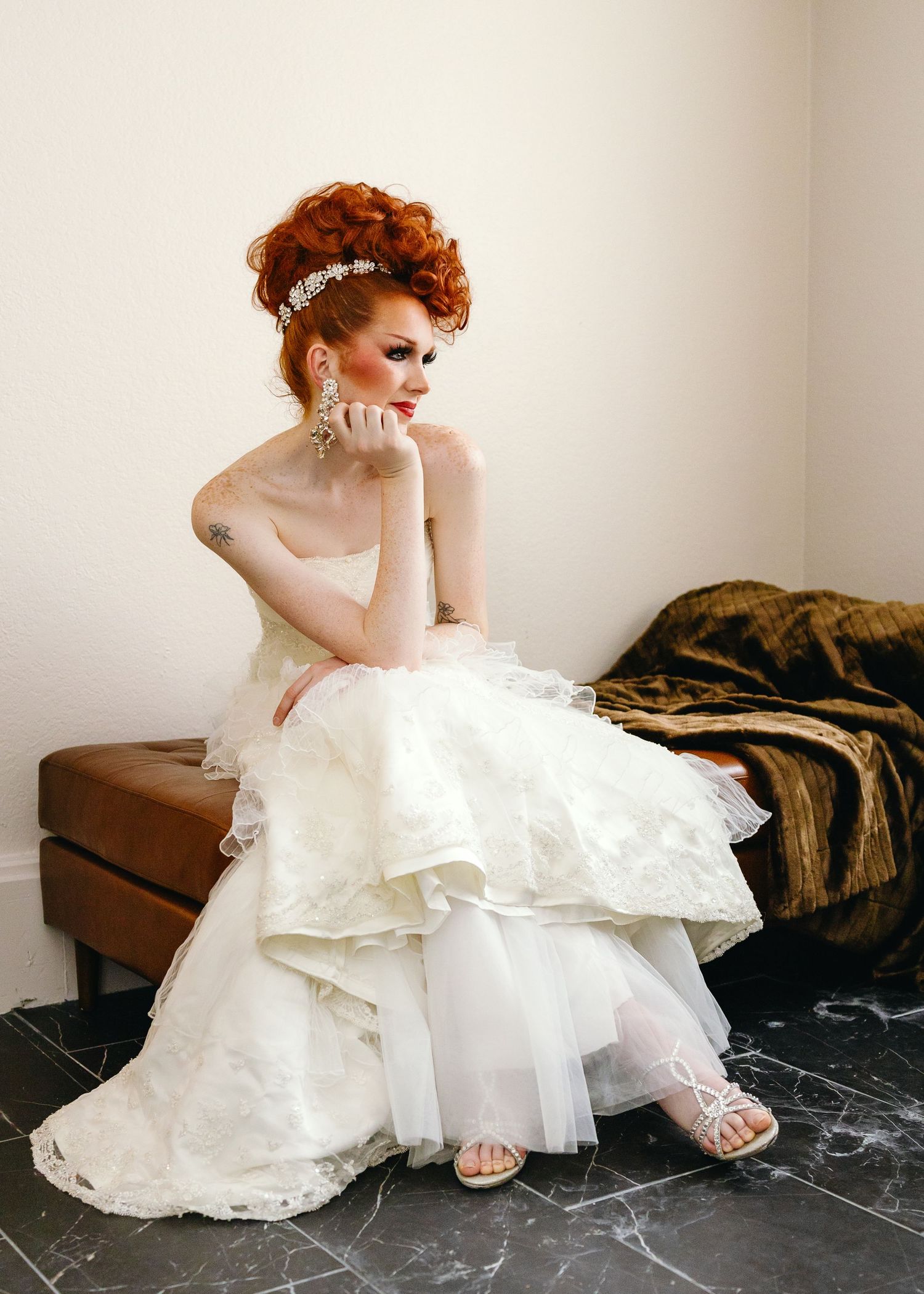 Elegant bride in layered white gown with red updo hairstyle, crystal accessories, seated pensively in a bridal suite.