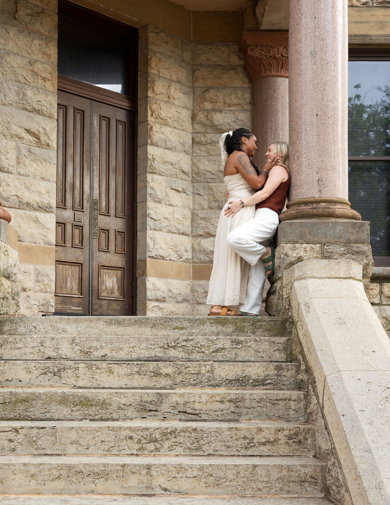 Two women share a romantic moment on stone steps outside a historic building with ornate columns and wooden doors.