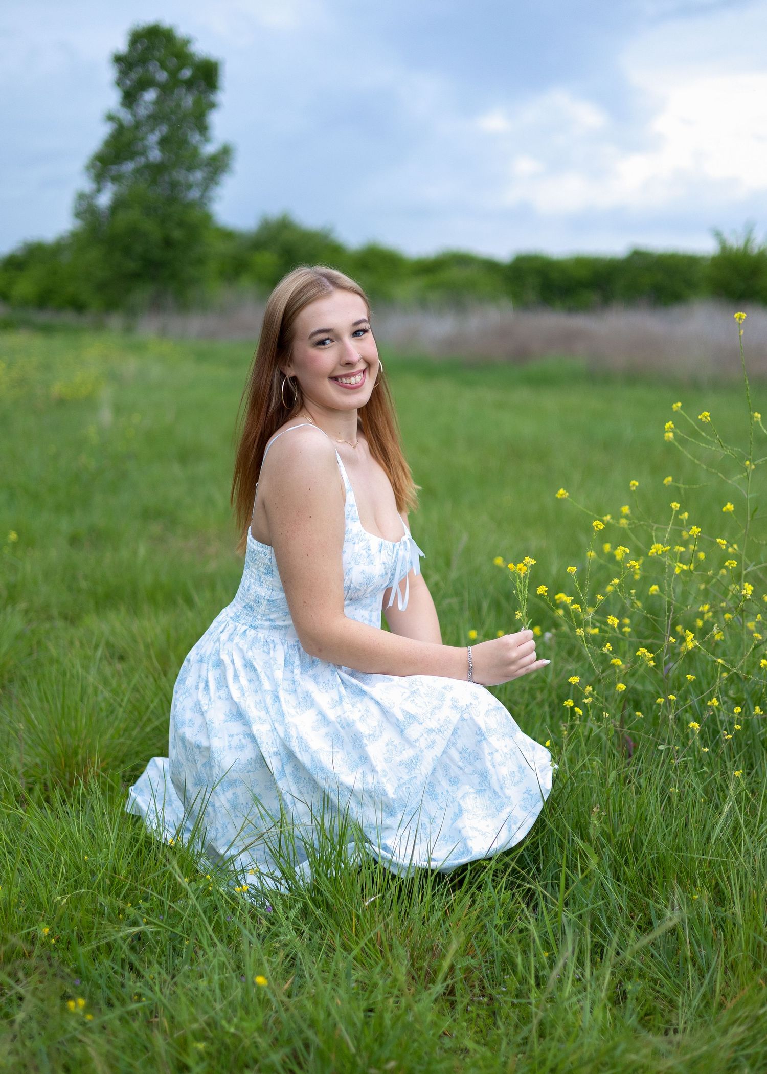 Young woman in a floral sundress smiling while sitting in a green meadow with yellow wildflowers on a cloudy day.