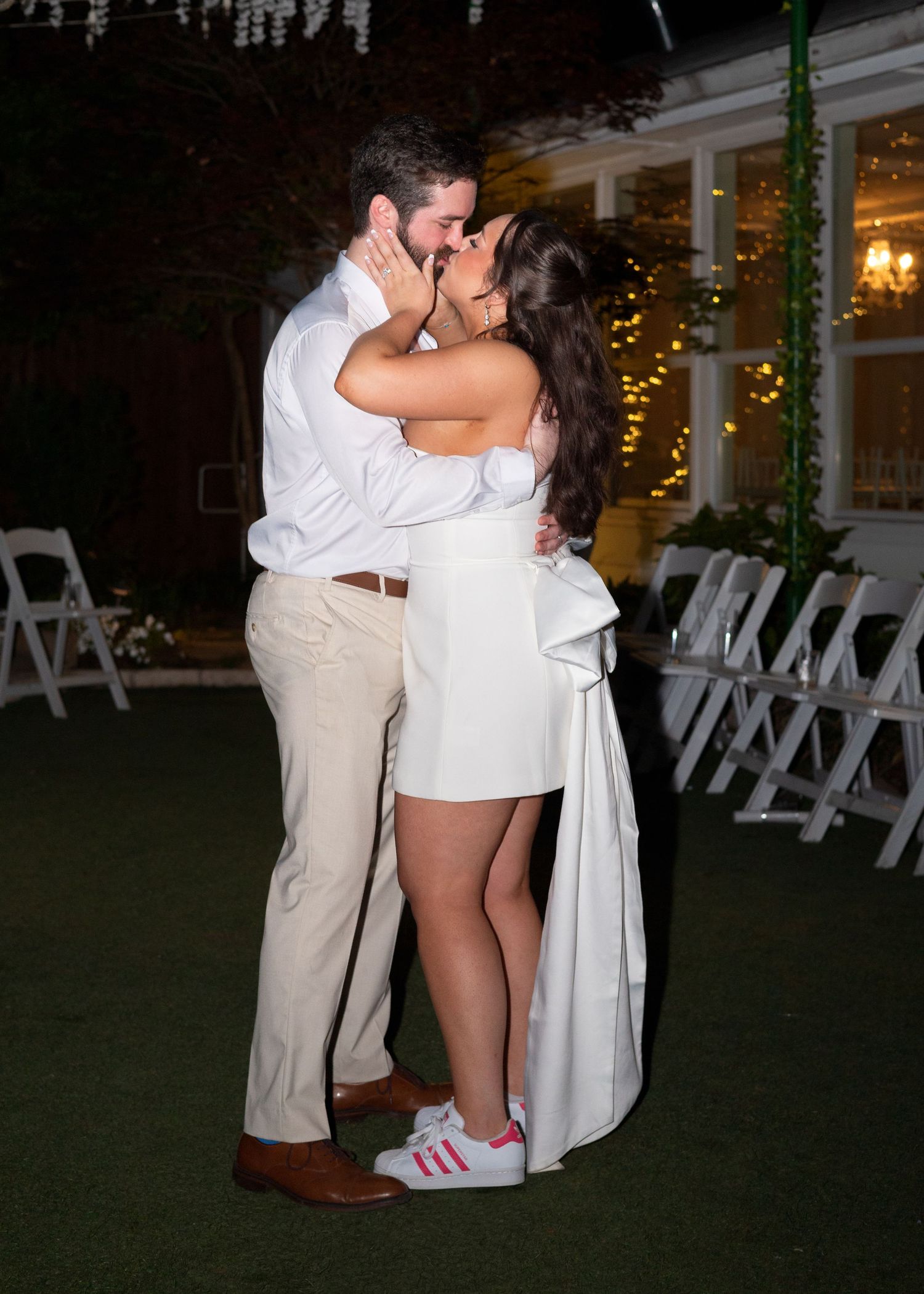 Couple sharing a romantic kiss in white outfits at an outdoor evening wedding reception with string lights.