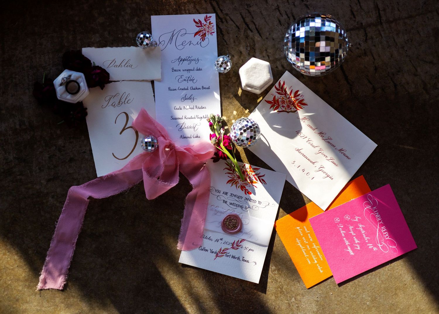 Elegant wedding stationery flat lay featuring table numbers, menus, and colorful invitation cards with pink ribbon and disco ball.