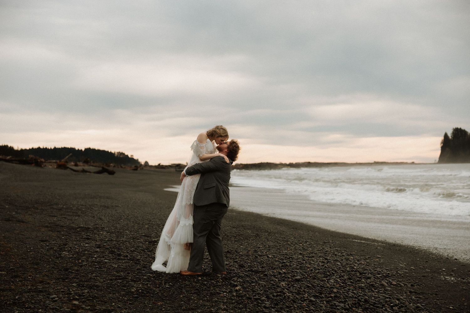Bride and groom sharing a romantic kiss on a dark sandy beach with ocean waves and cloudy sky behind them.