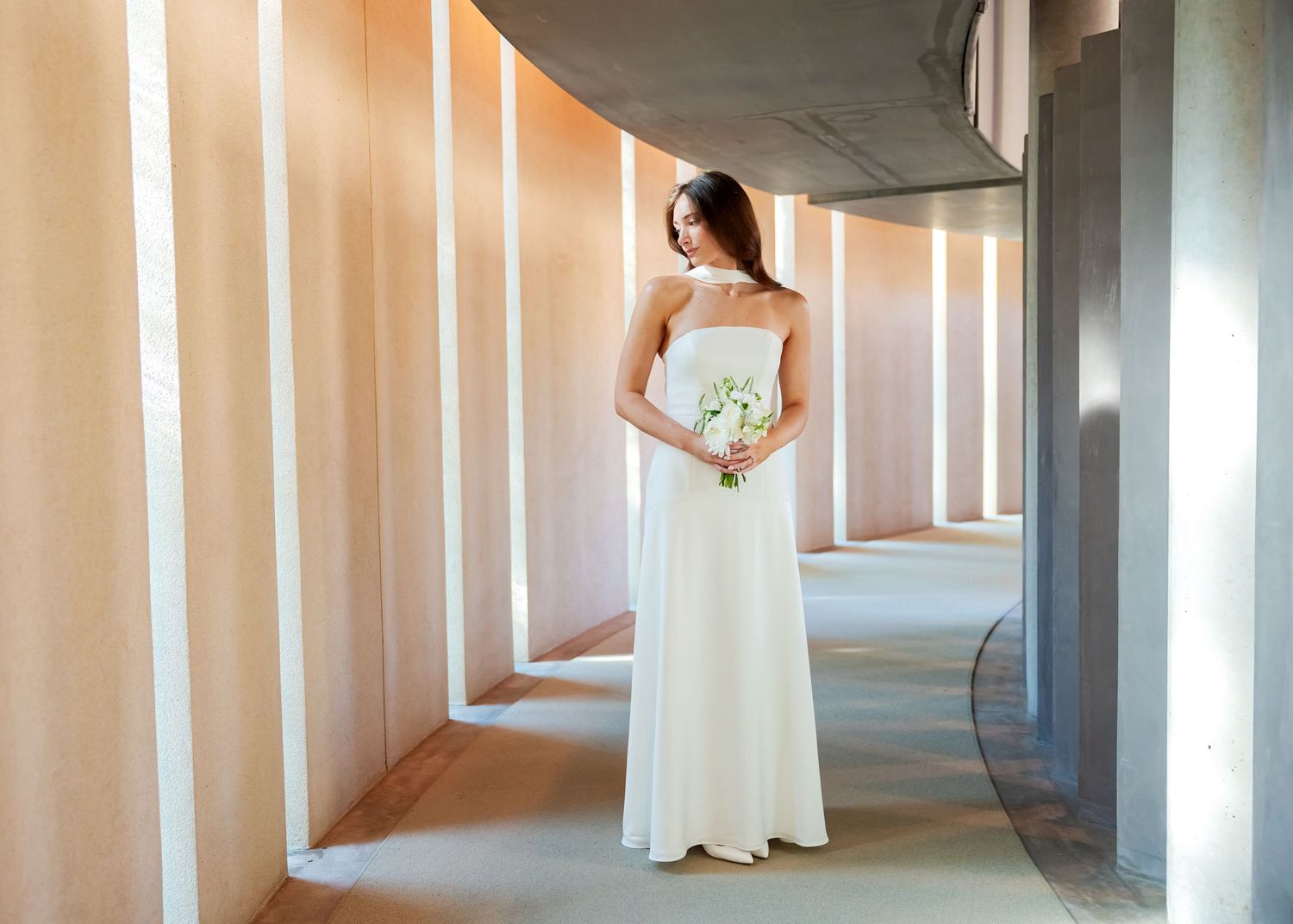 Bride in white strapless gown holding bouquet stands in elegant corridor with warm peach walls and natural light.