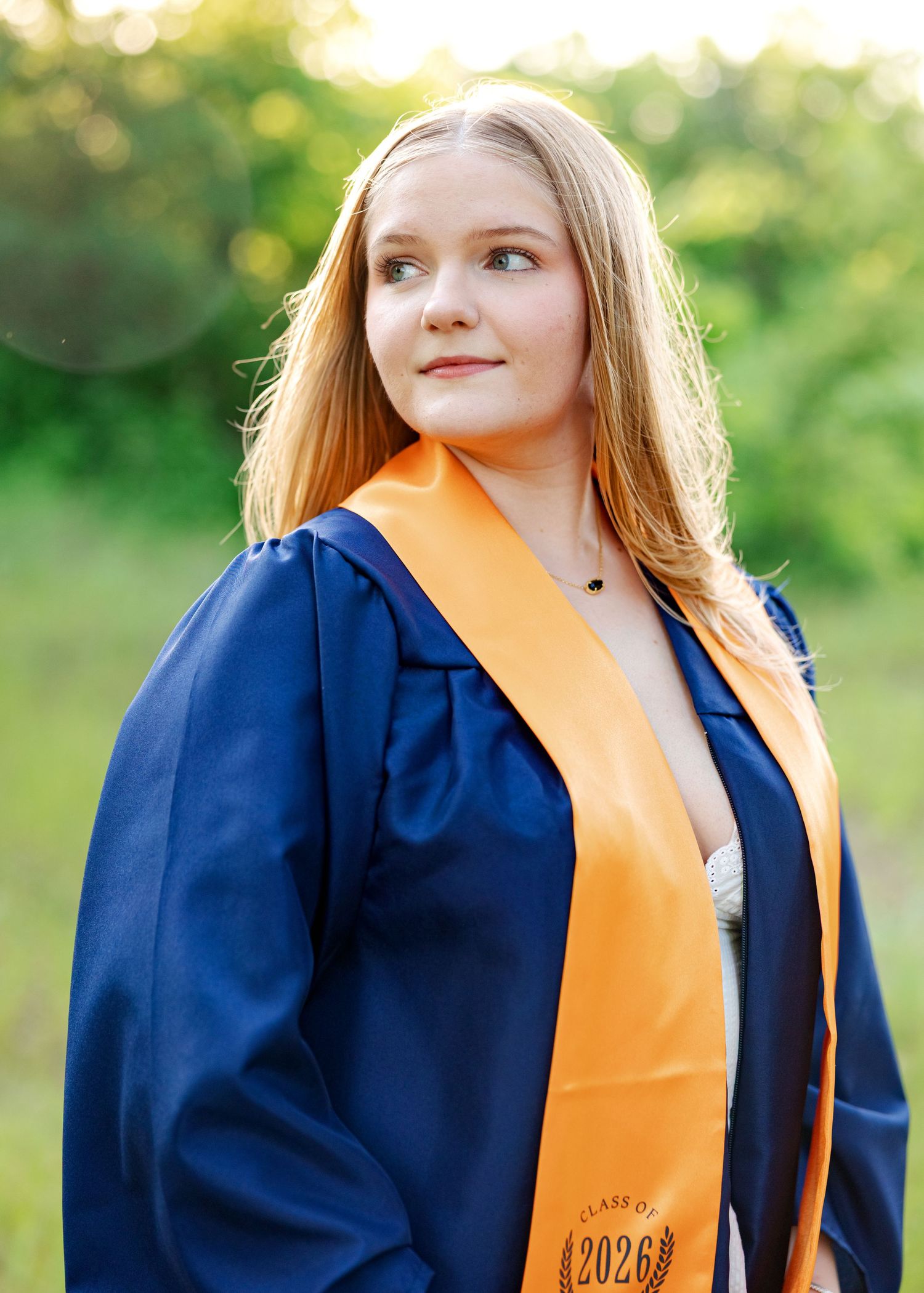Young woman in navy blue graduation gown with orange Class of 2026 sash posing outdoors with green foliage background.