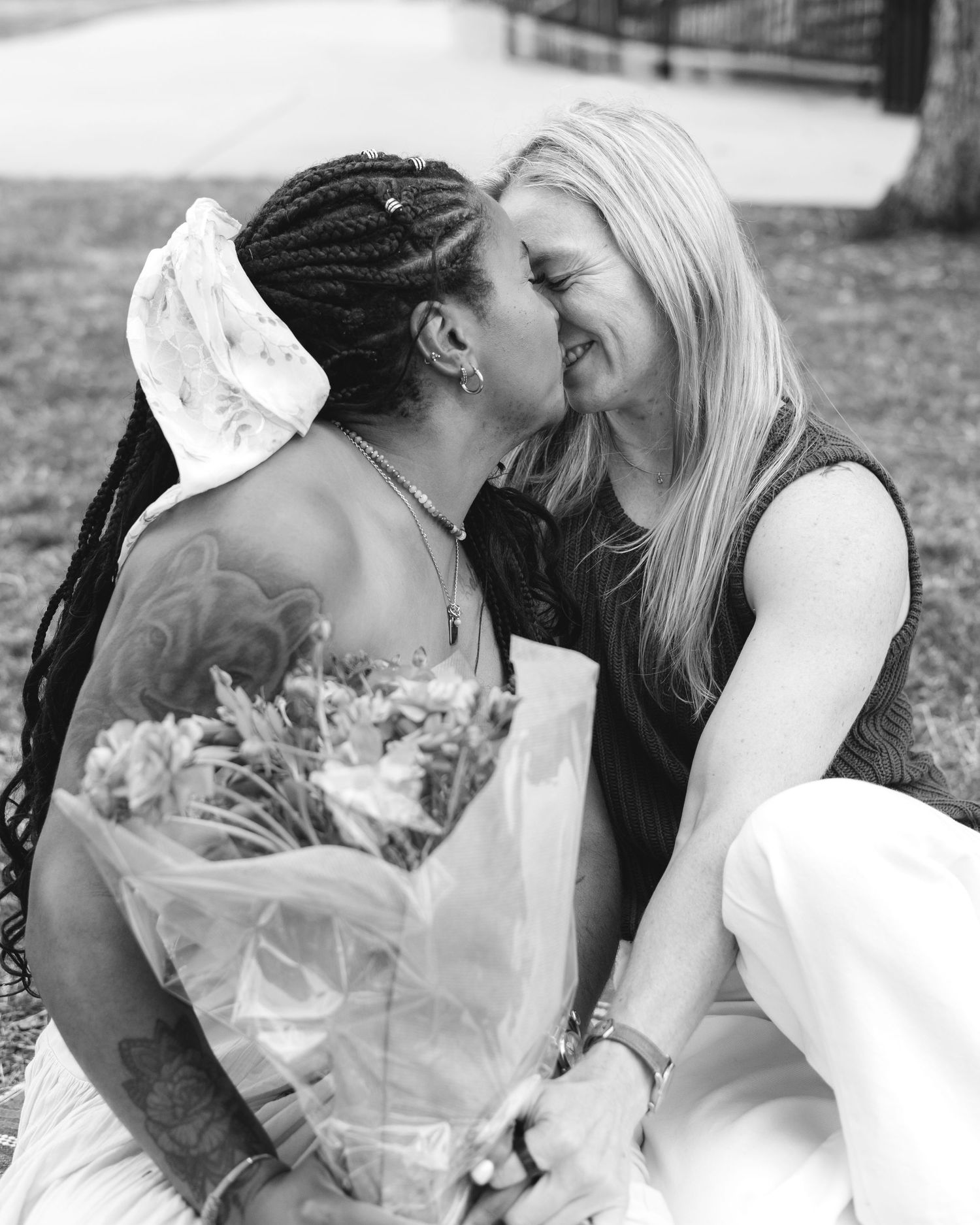 Black and white photo of two women sharing a kiss, one holding a bouquet, dressed in white for a wedding celebration.