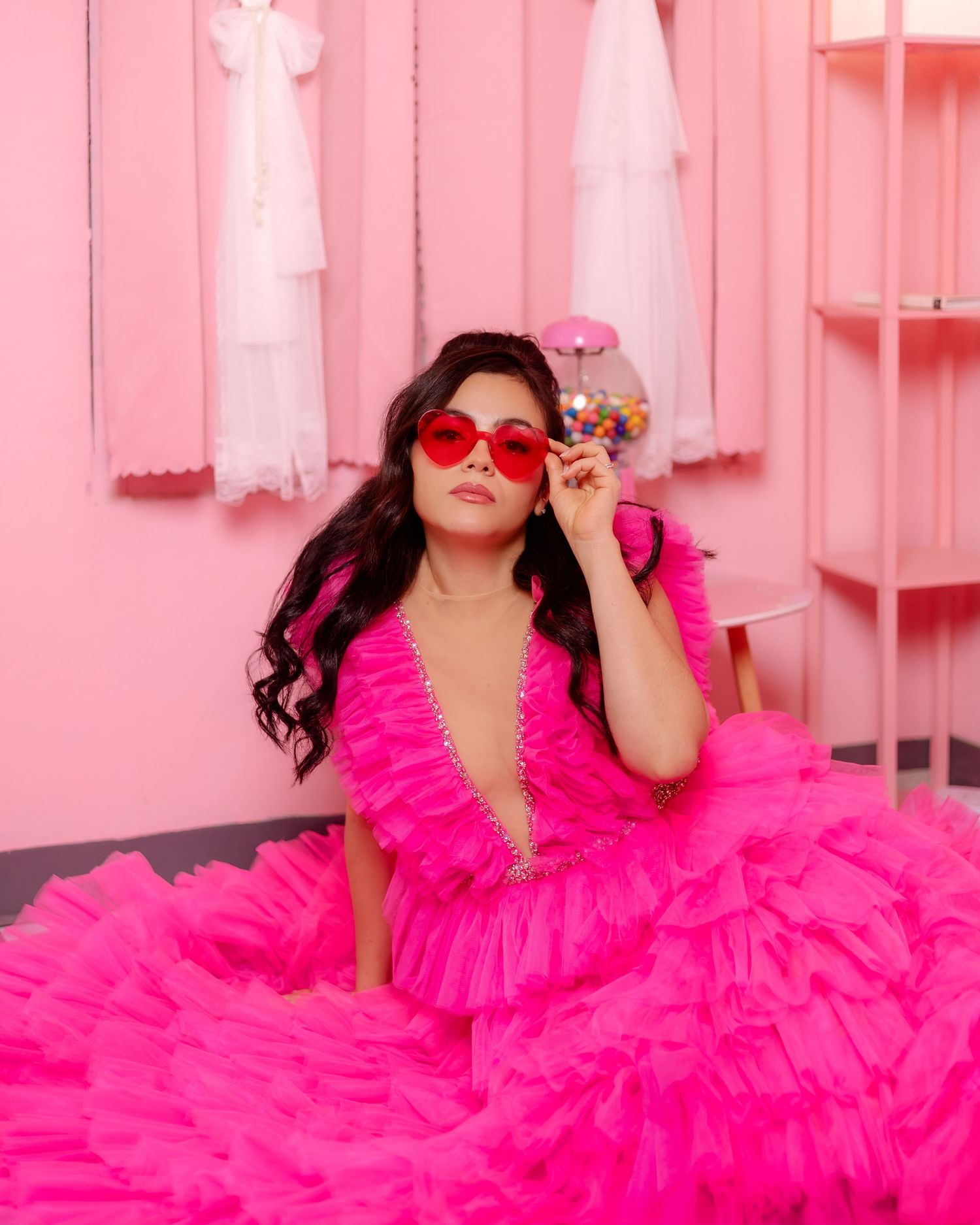 A bright hot pink tulle gown with ruffles and deep v-neckline photographed against a pink wall with white decorations.
