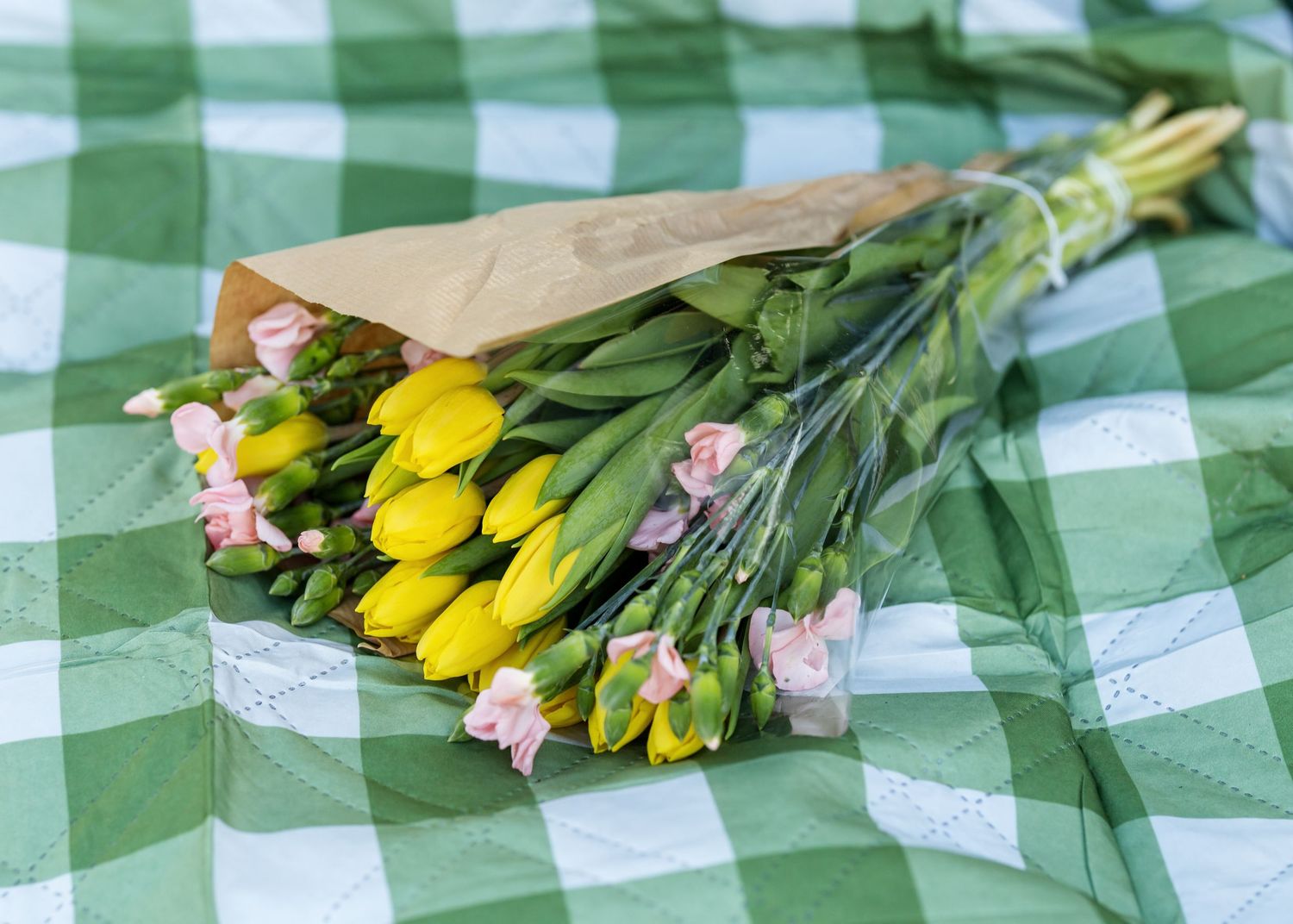 Yellow tulips and pink flowers arranged on green checkered picnic blanket.