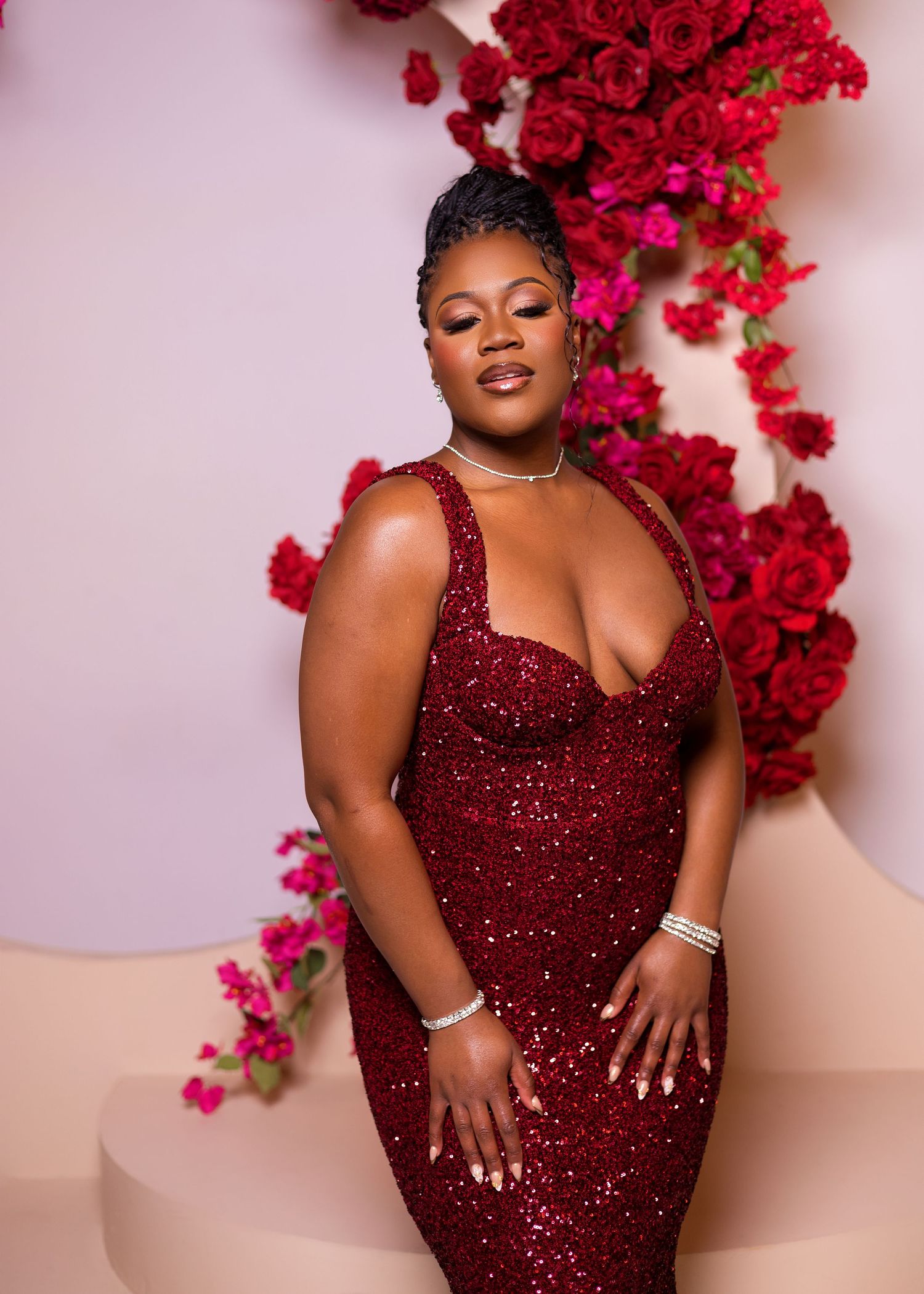 Model in sparkling burgundy evening gown poses against floral backdrop with red roses.