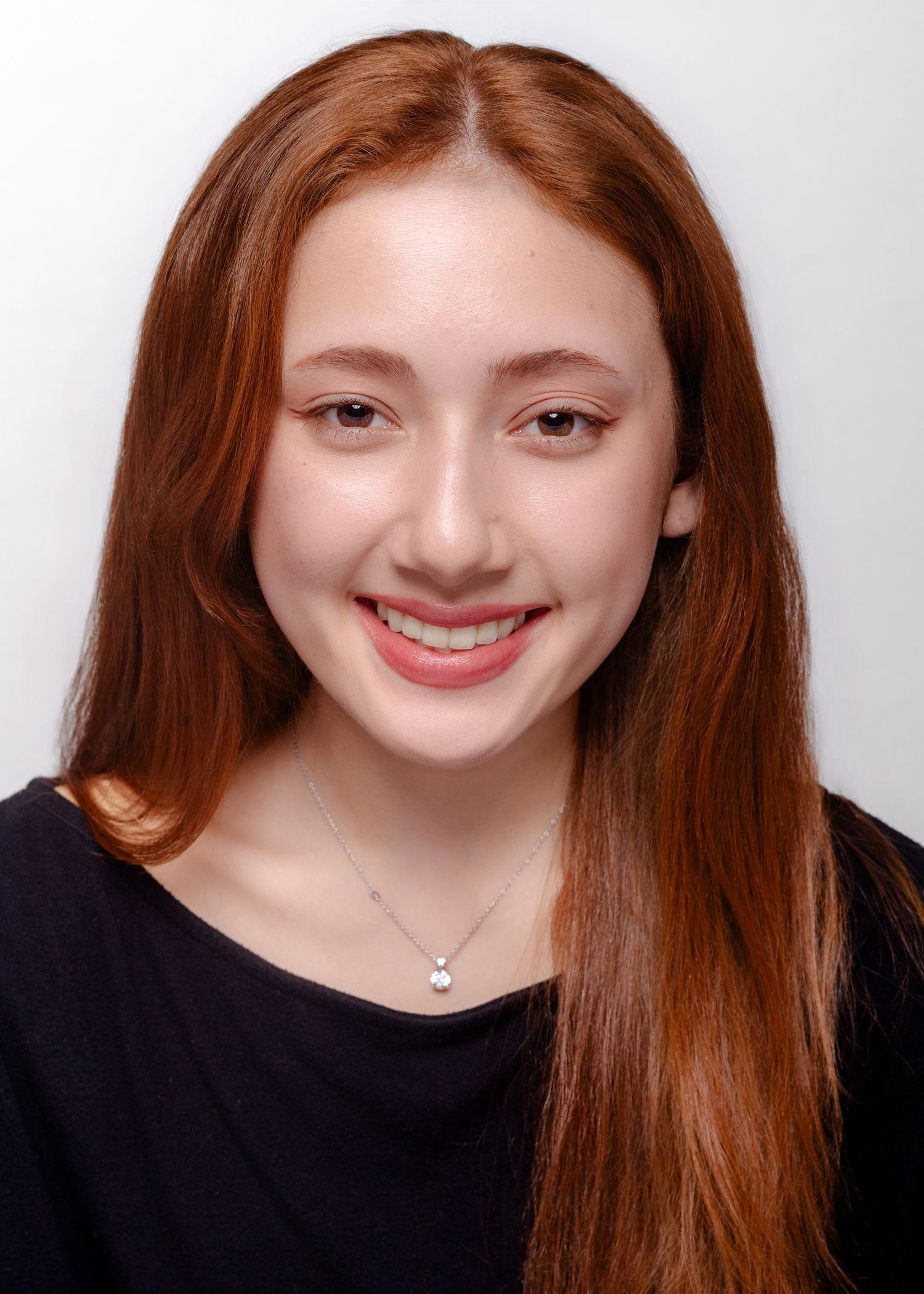 Portrait of a smiling young woman with long auburn hair wearing a black top and delicate necklace against a light background.