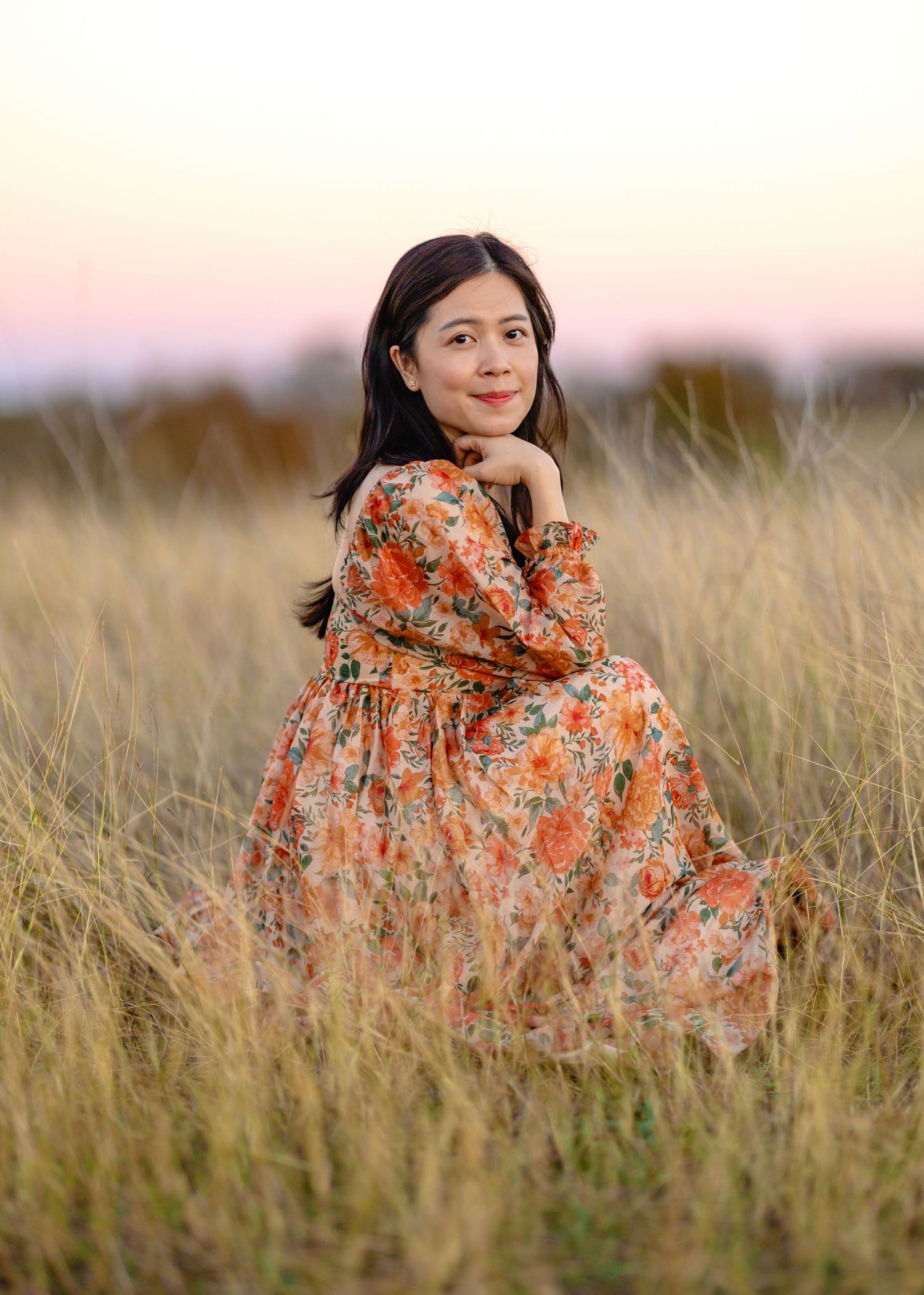 Woman in floral orange dress poses gracefully in tall grass field during sunset.