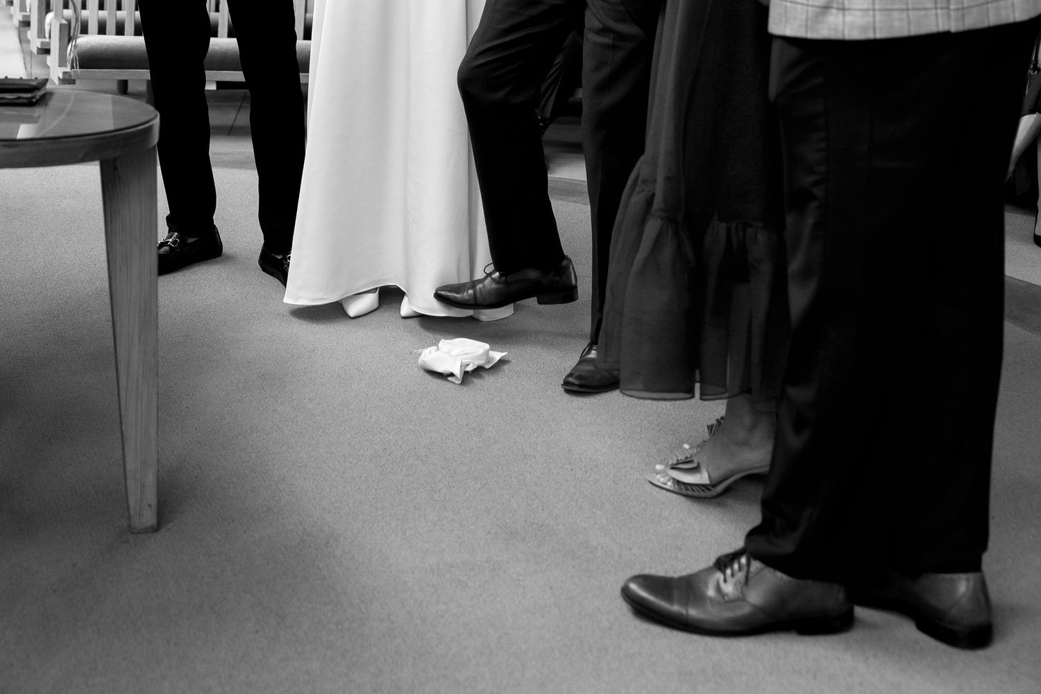 Black and white photo of wedding guests standing in a circle, showing formal shoes and a white bridal gown hem.