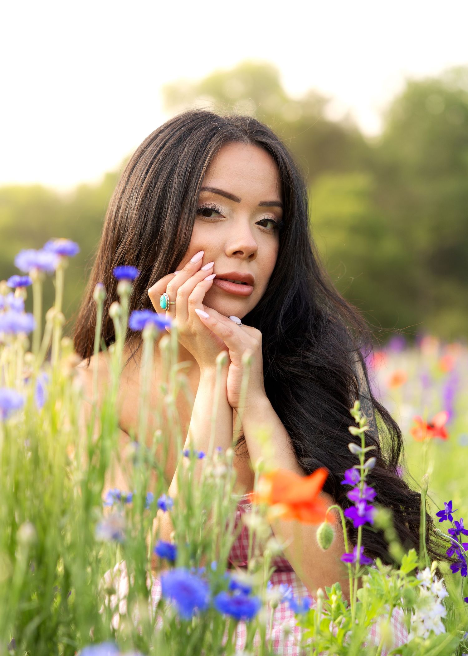 Young woman with long dark hair poses among colorful wildflowers, resting chin on hands in a sunlit field.