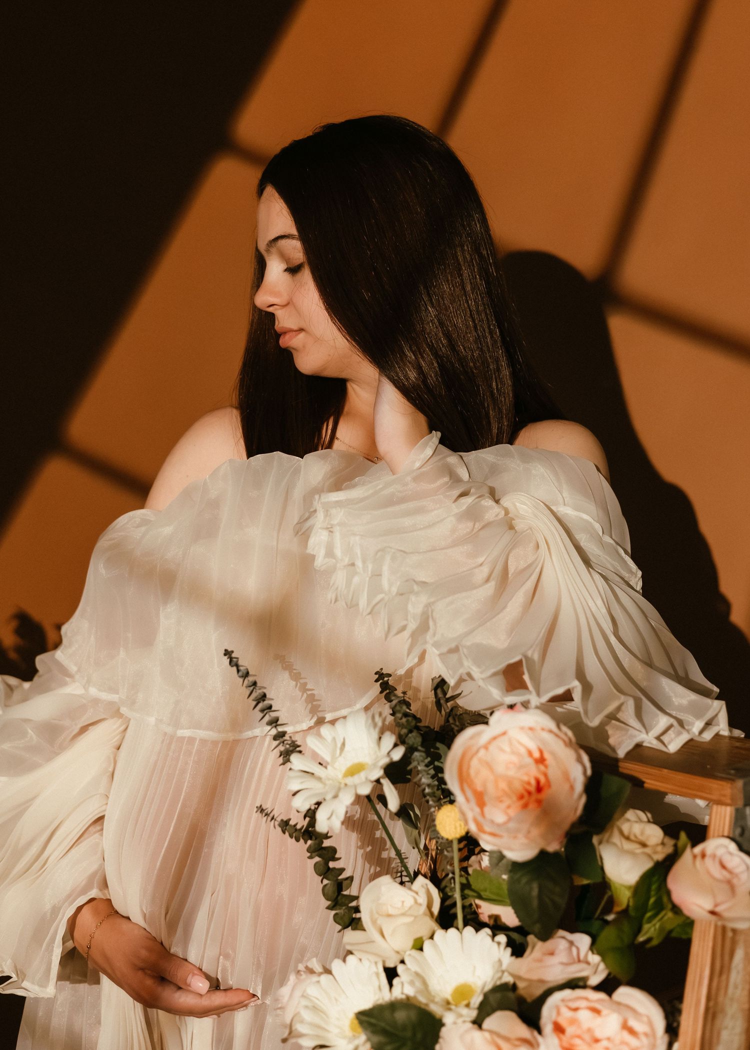 Pregnant woman in white ruffled gown poses with blush roses and daisies in warm studio light.