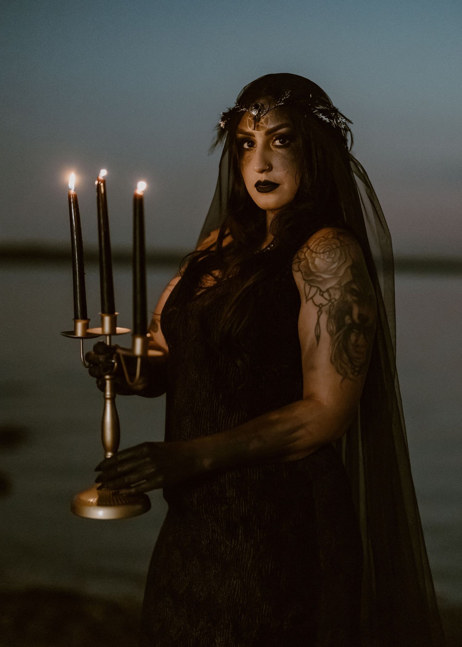 A figure in a black dress and veil holds a candelabra against a moody twilight backdrop near water.