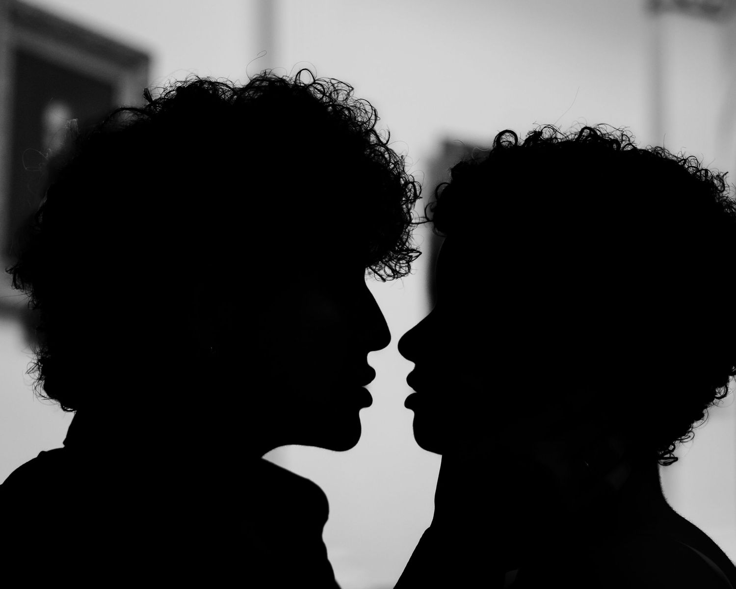 Black and white silhouette of two people with curly hair facing each other closely, almost kissing.