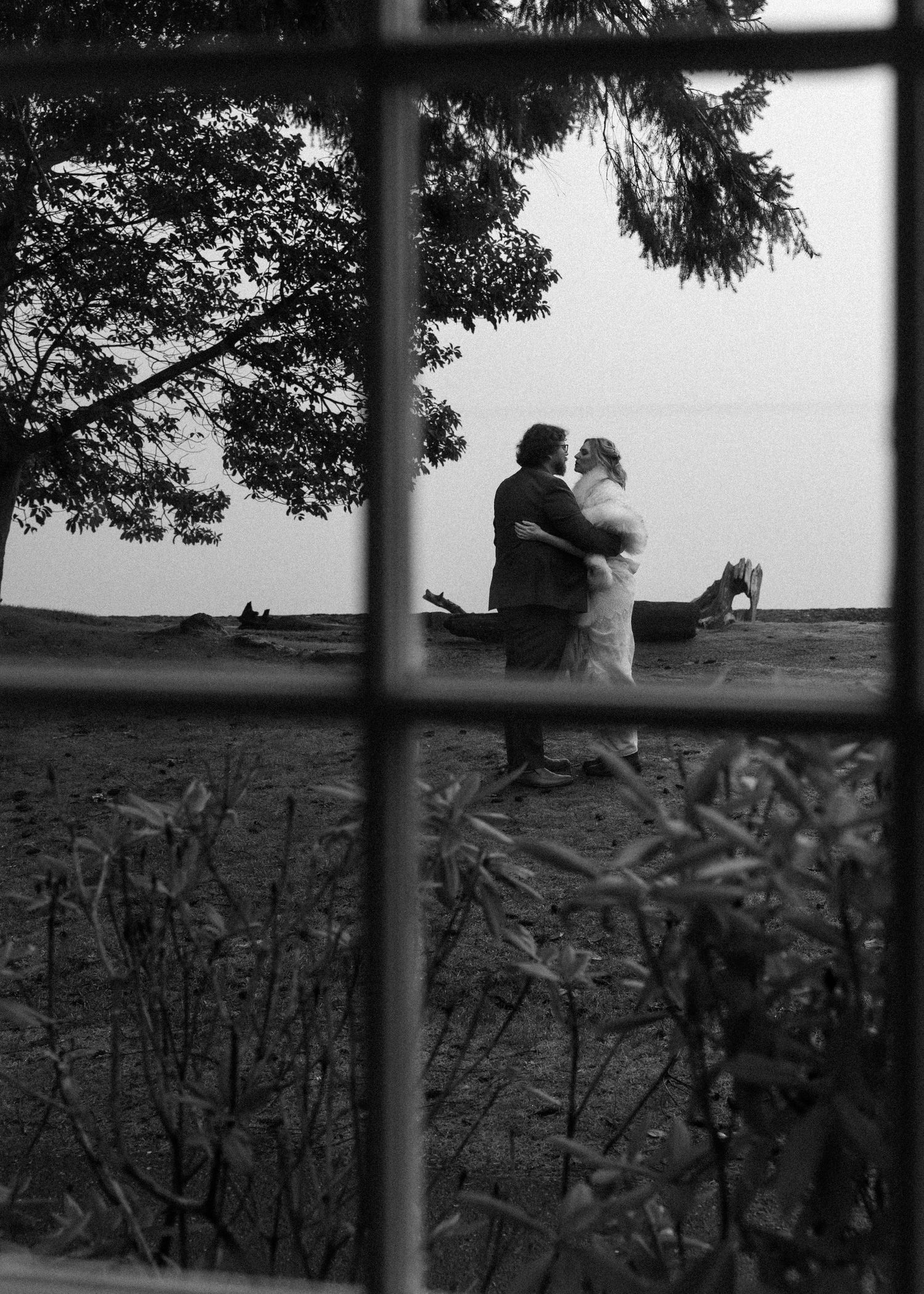 Black and white photo of a couple embracing on a beach, viewed through a window frame with trees overhead.